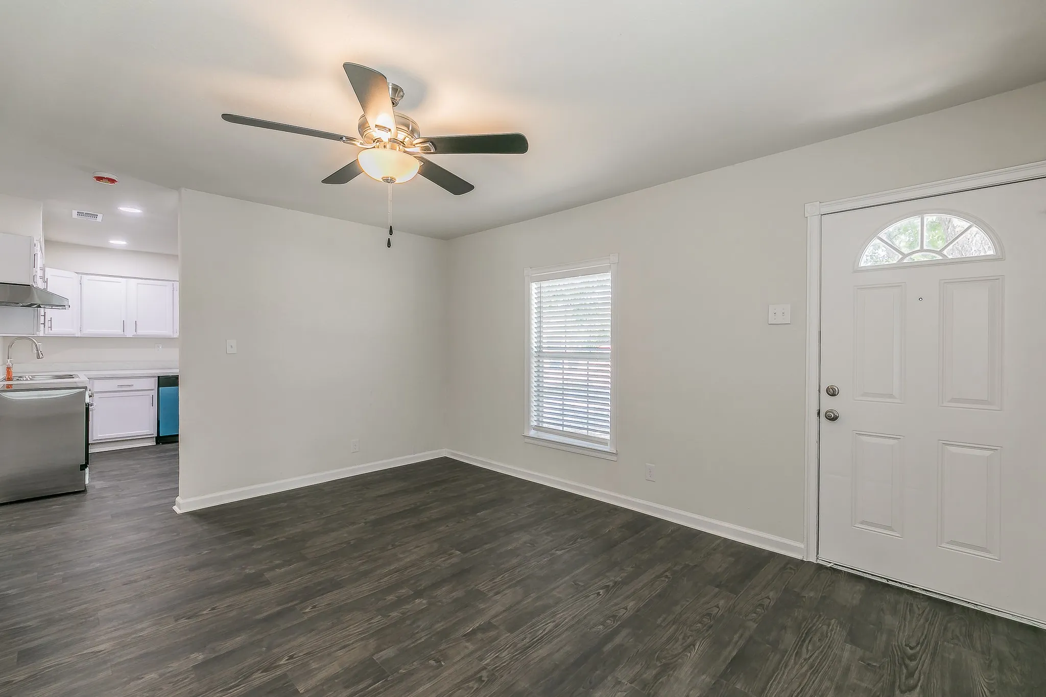 Wood-finish flooring throughout, ceiling fan with integrated lighting, white paneled door with arched transom, and a window with horizontal blinds