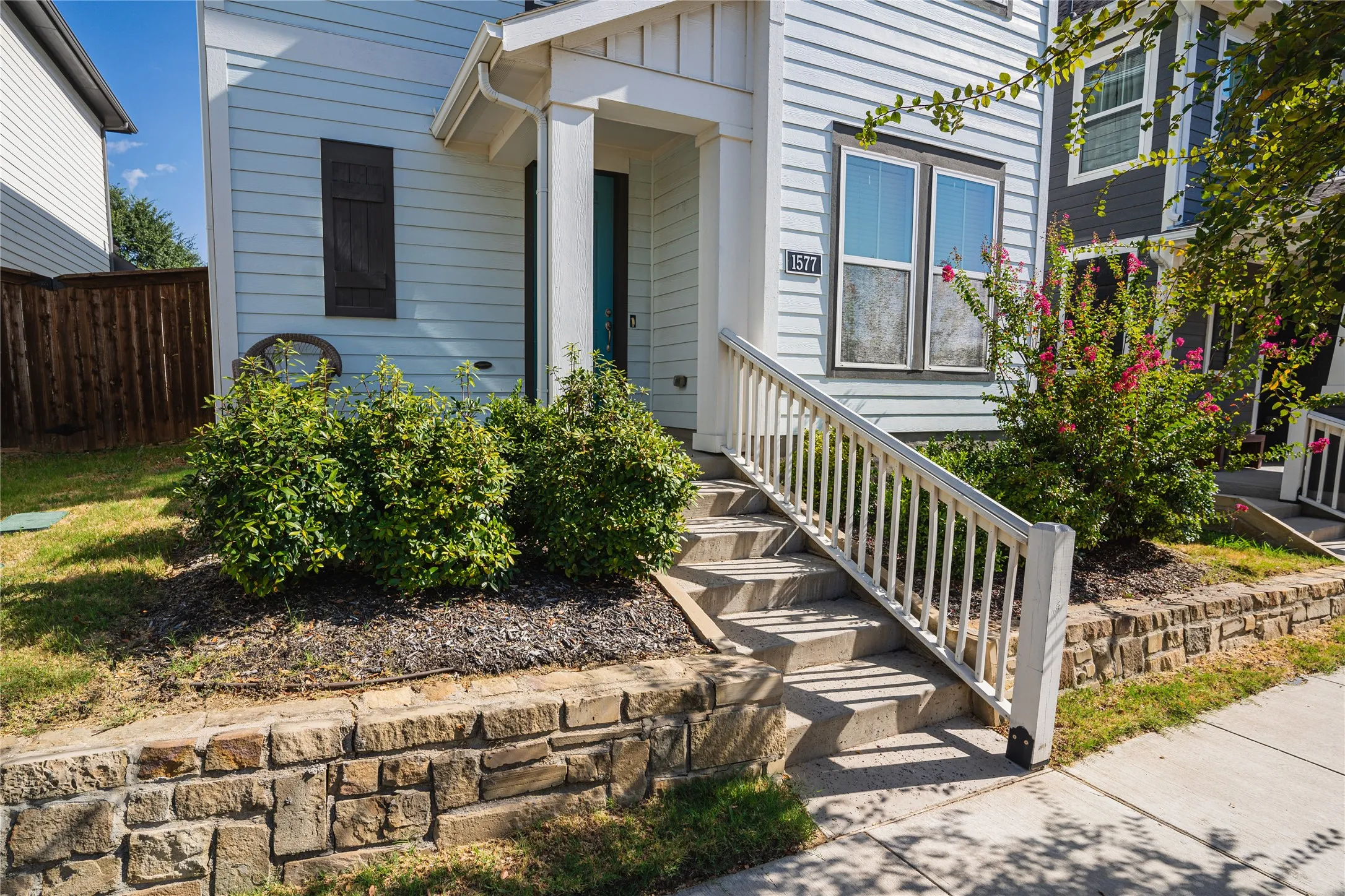Stone retaining wall, concrete steps with white railing, light-colored siding, dark shutters, and established landscaping