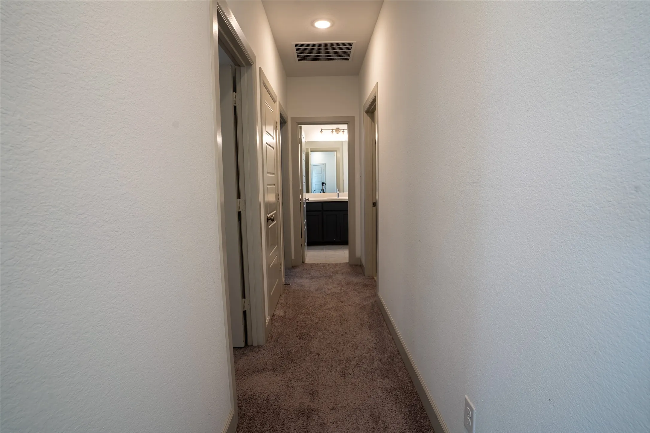 Recessed lighting illuminates a hallway with neutral carpet flooring, leading to a bathroom featuring a dark wood-finish vanity