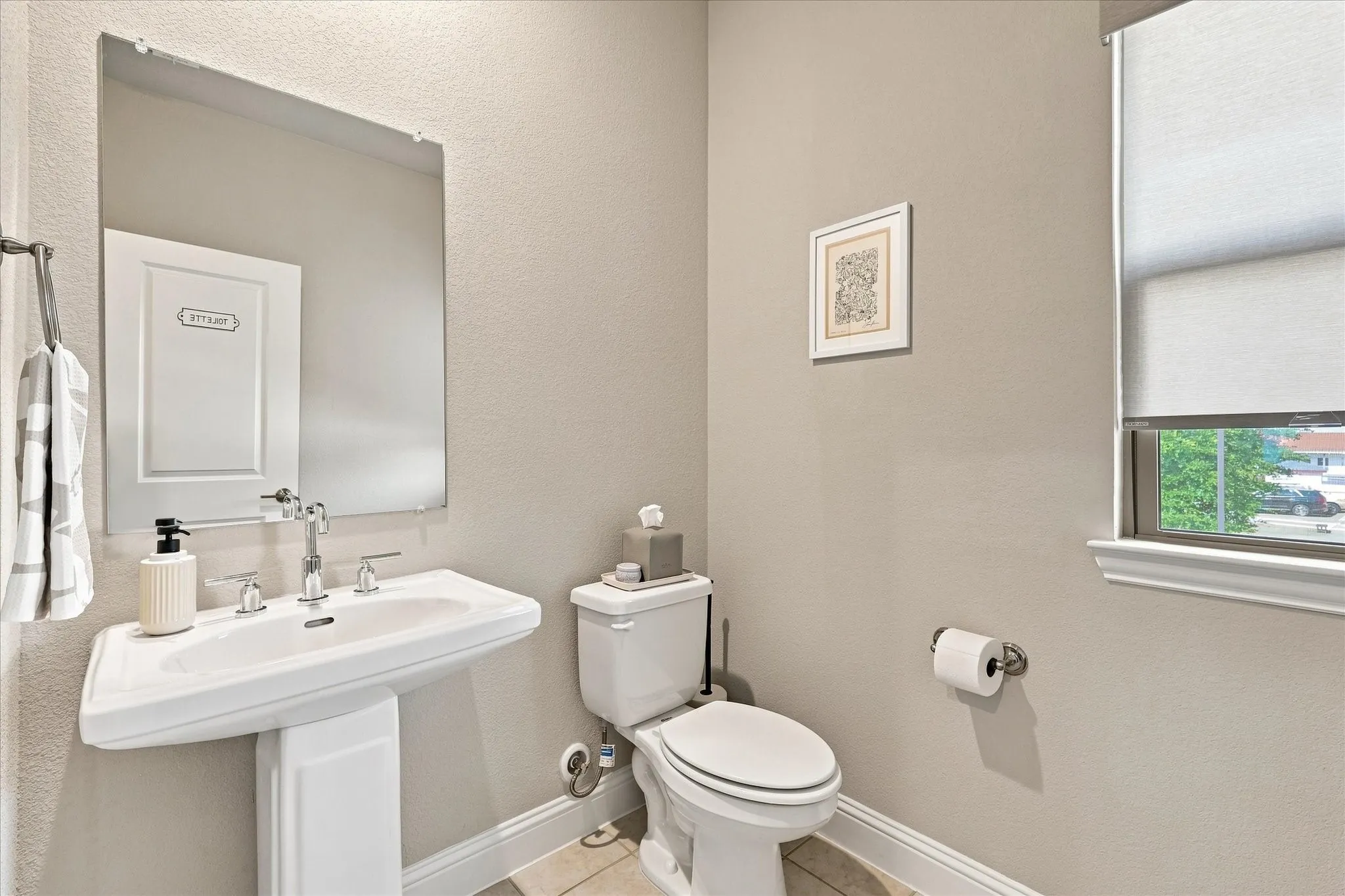 Pedestal sink with chrome fixtures, wall-mounted mirror, commode, and a window with roller shade