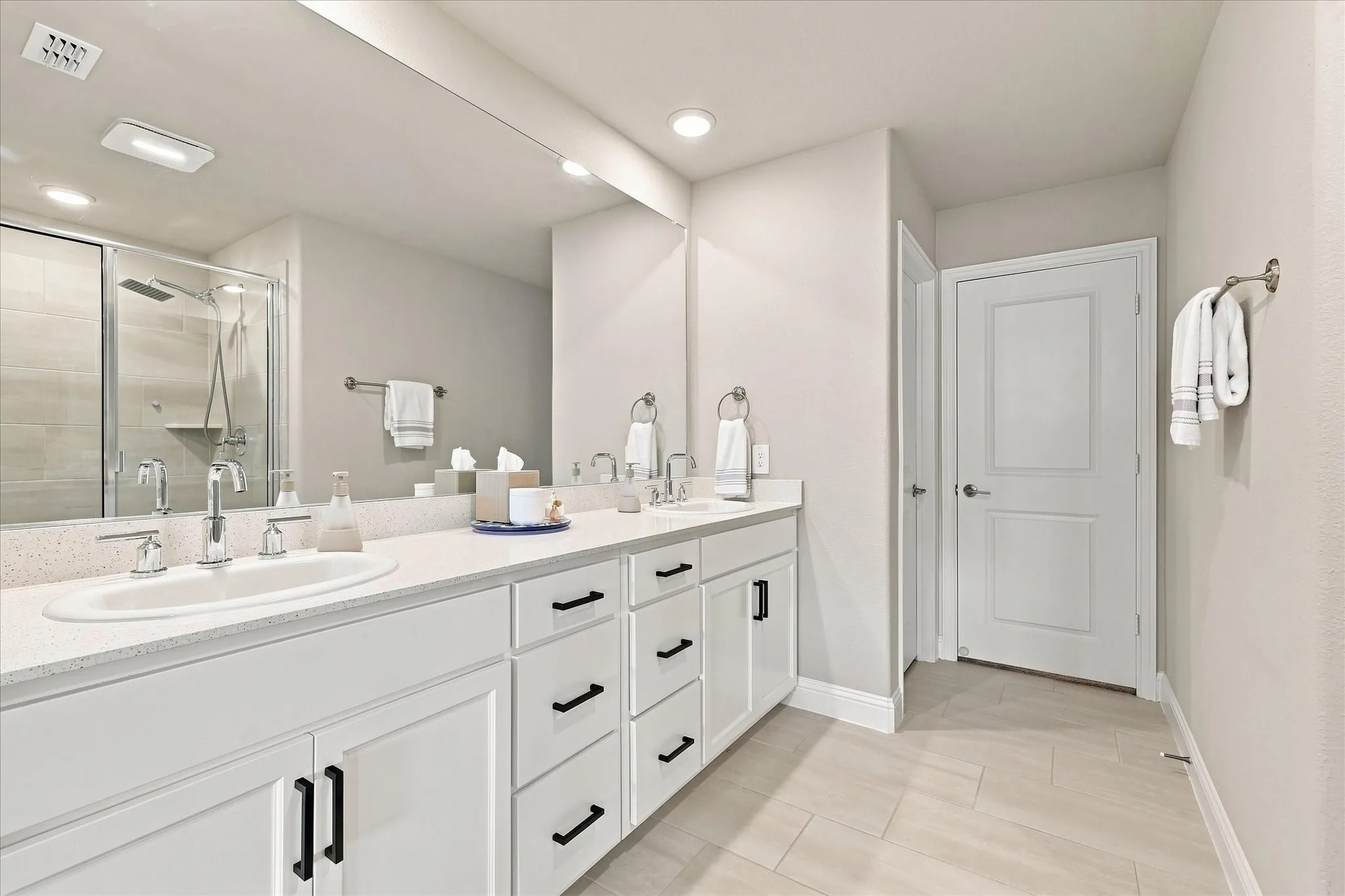 Dual undermount sinks with chrome fixtures, white vanity cabinetry with matte black hardware, expansive vanity mirror, glass-enclosed shower with tiled walls, and light-toned tile flooring