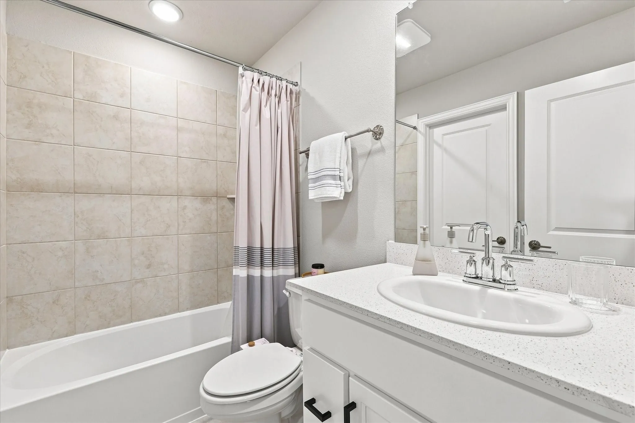 White vanity with a speckled countertop, a chrome faucet, a rectangular mirror, and a tub with light-colored wall tiling