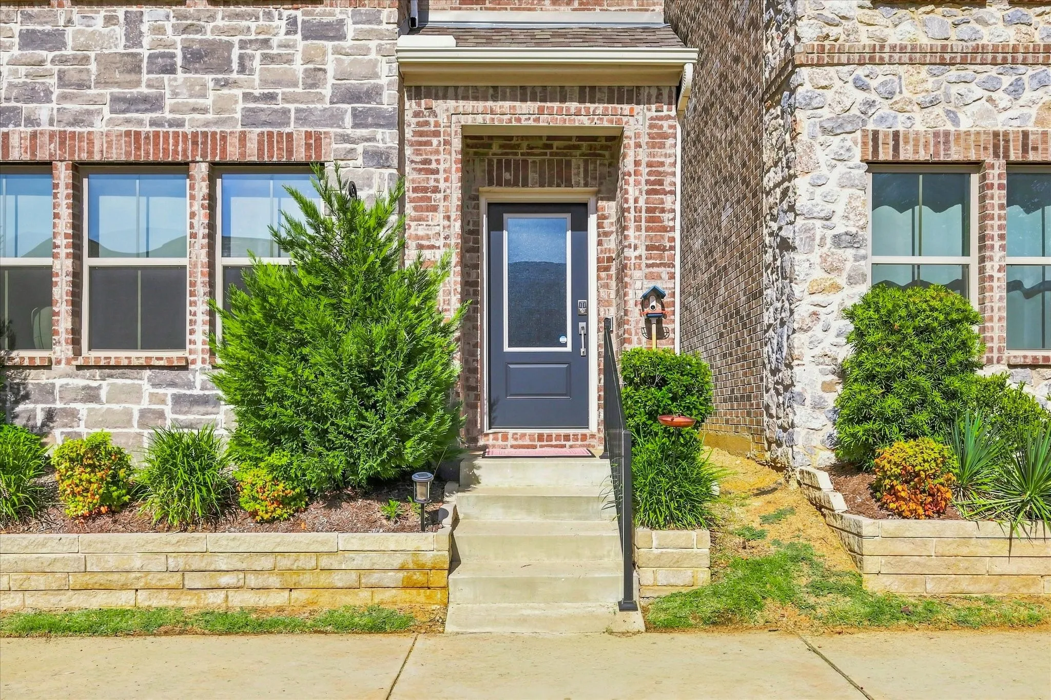Brick and stone facade, concrete steps, wrought iron handrail, dark paneled entry door with glass insert, and foundational landscaping