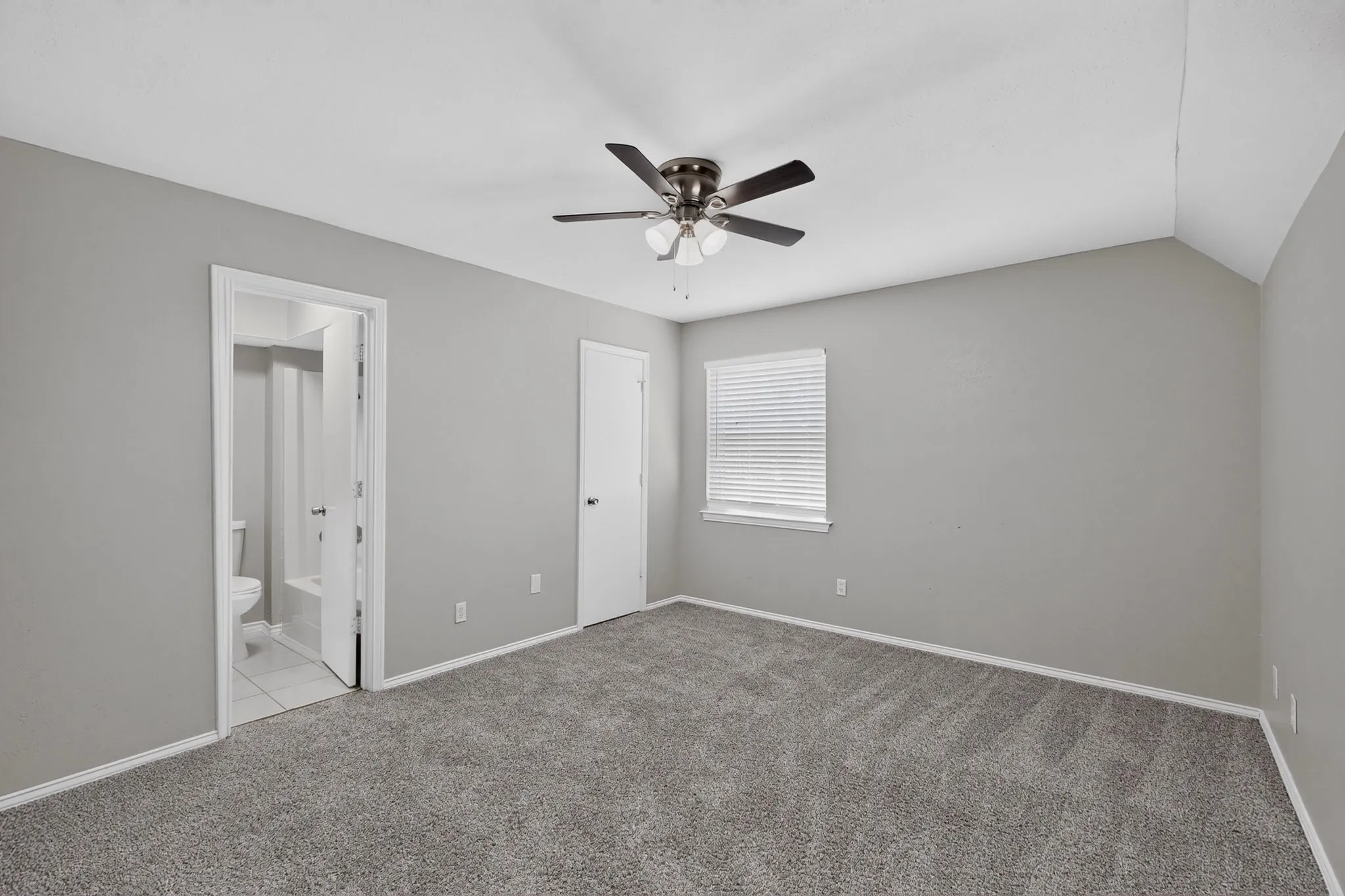 Carpeted flooring, vaulted ceiling, ceiling fan, window with blinds, and an attached bathroom with a toilet and tub