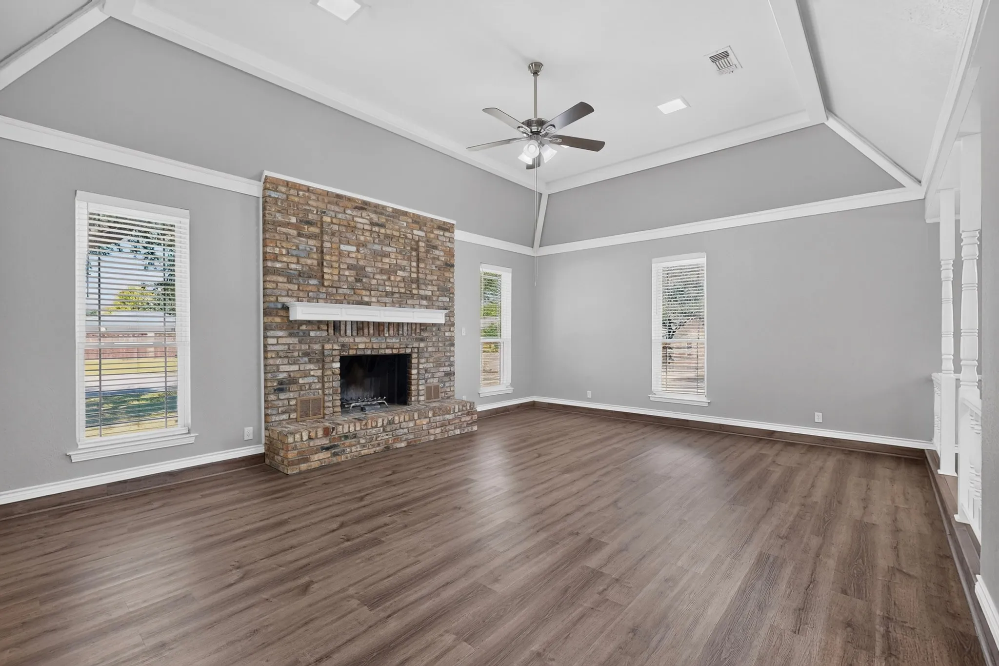 Vaulted ceiling, brick fireplace with a white mantel, ceiling fan, multiple windows, and wood-finish flooring