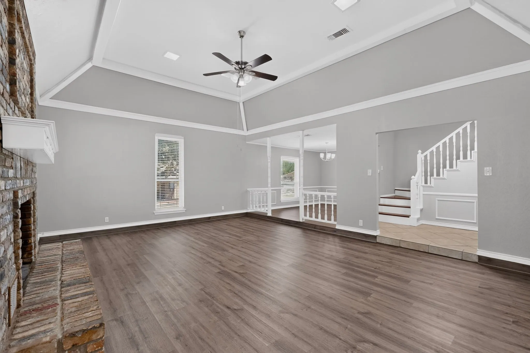 Spacious room featuring vaulted ceilings, a prominent brick fireplace with a white mantel, wood-finish flooring, and a staircase with white balusters