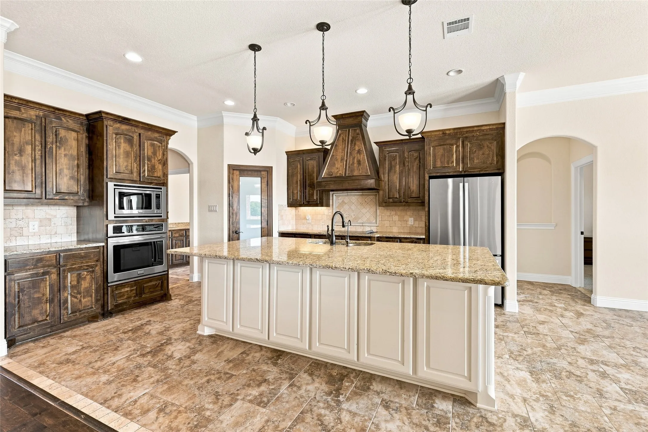 Gourmet kitchen featuring a large central island with light-colored cabinetry, stainless steel appliances, dark wood-finish cabinetry, tile flooring, and decorative pendant lighting