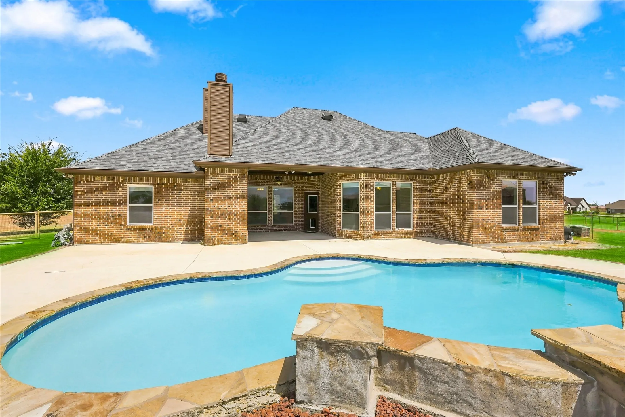 Brick exterior with a shingled roof, a chimney, multiple windows, a concrete patio, and an in-ground swimming pool with stone coping