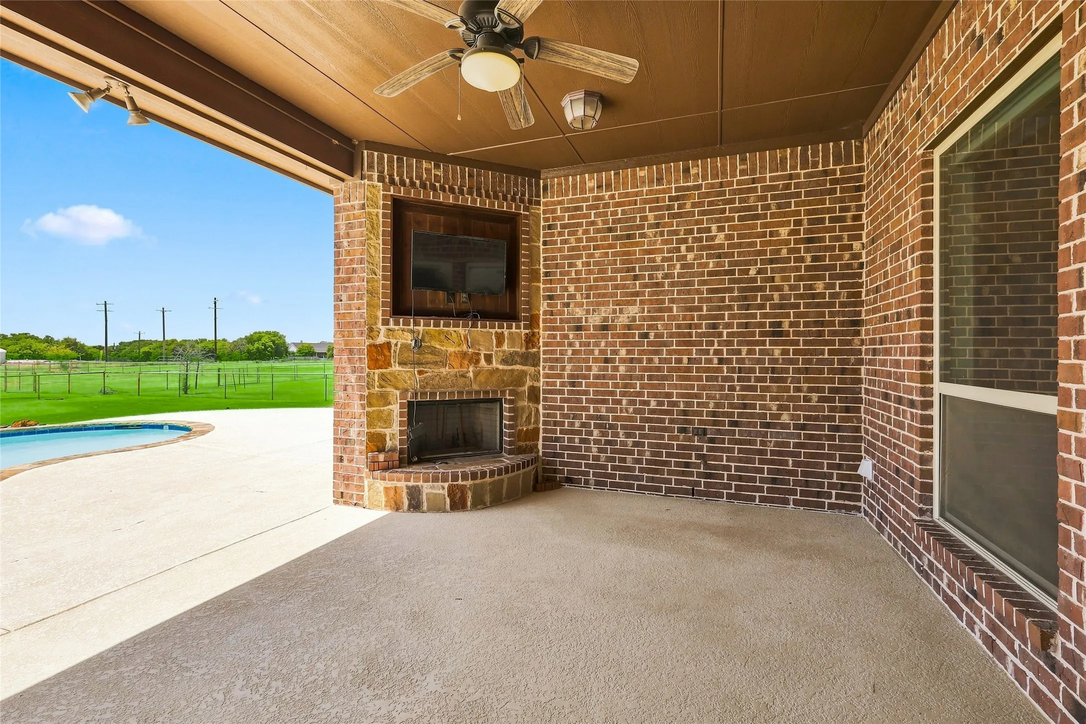 Covered outdoor patio with a built-in fireplace, brick and stone accents, ceiling fan, and adjacent swimming pool