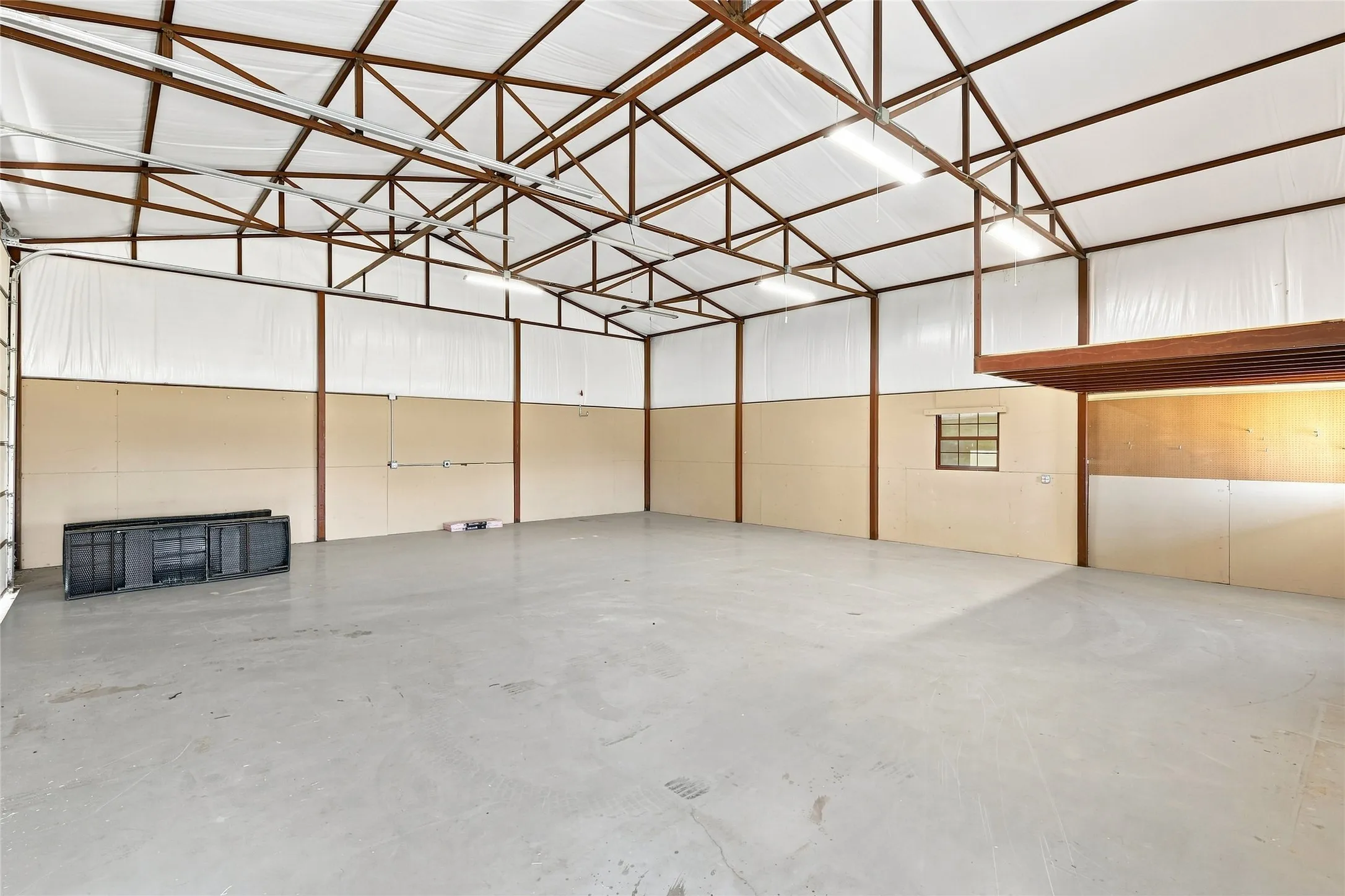 Expansive concrete flooring, high vaulted ceiling with exposed metal truss work, upper level storage platform, and multiple fluorescent light fixtures