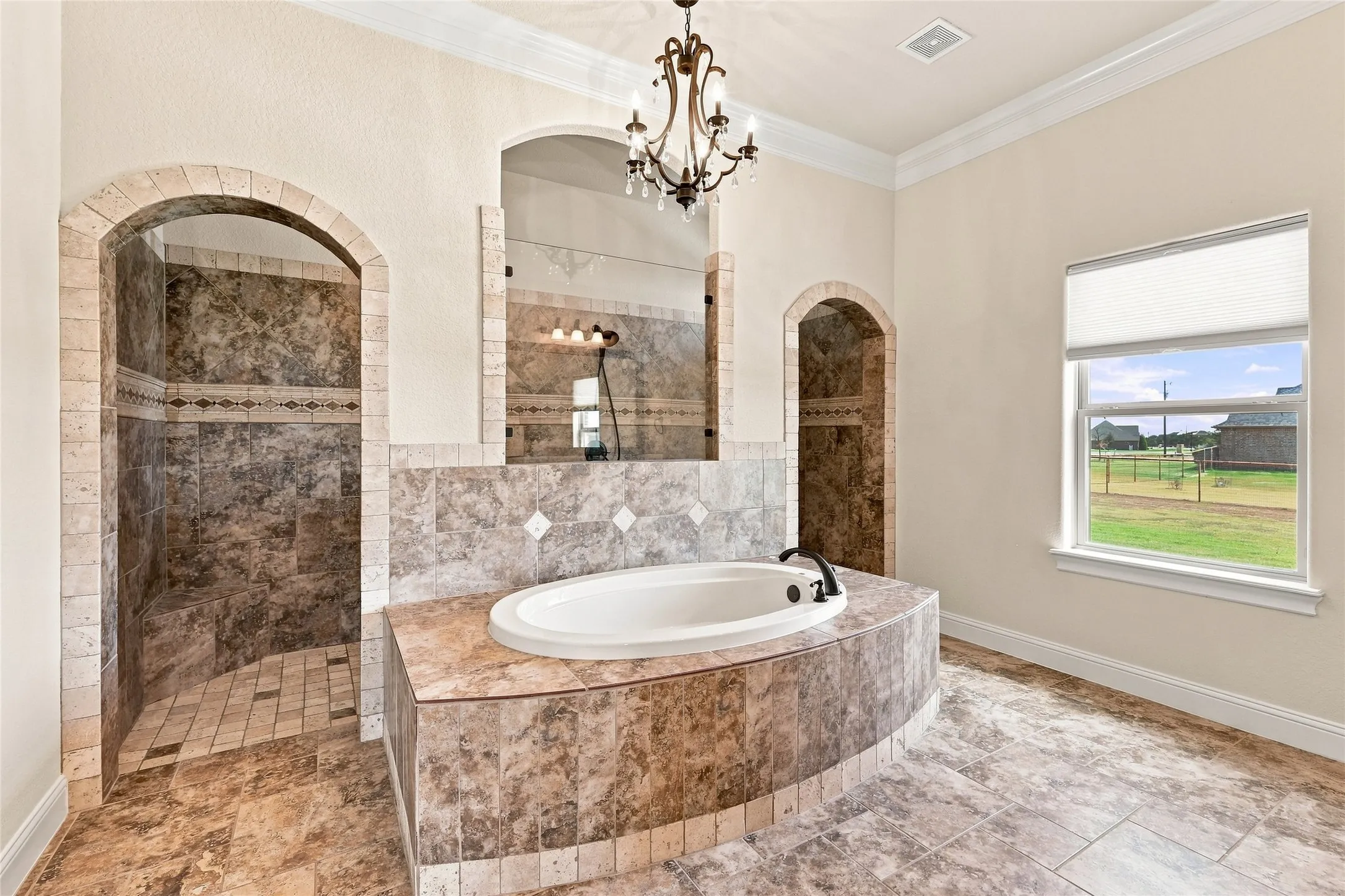 Spacious bathroom featuring an oval soaking tub, two arched walk-in showers, decorative floor tiling, and an ornate chandelier