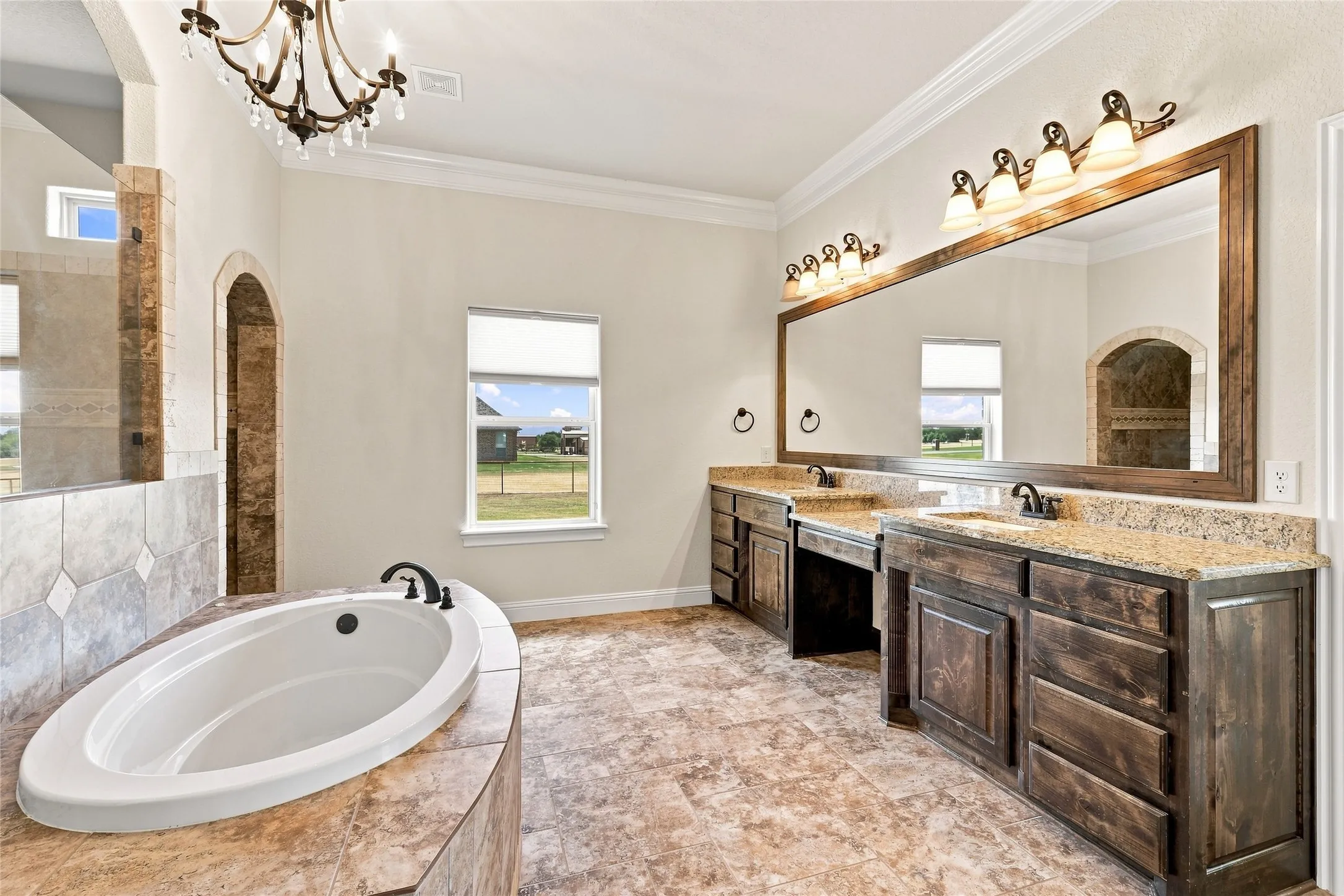 Dual wood-finish vanities with light-colored countertops, a built-in soaking tub, and a decorative chandelier