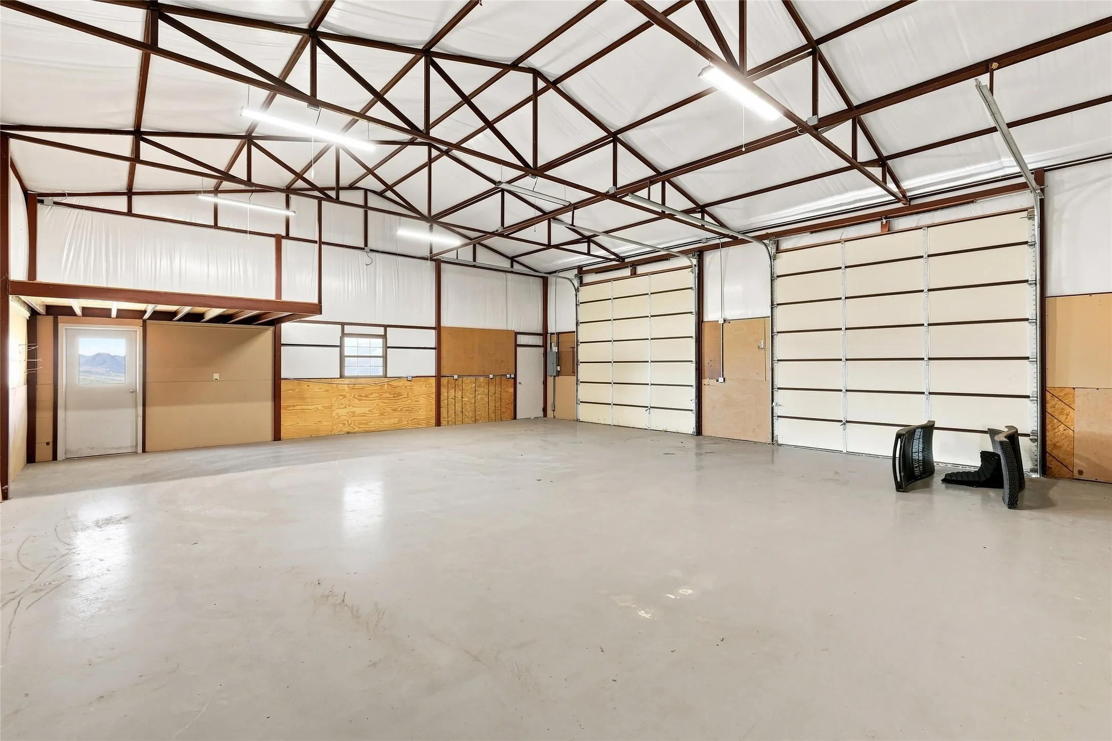 Expansive open industrial space with a concrete floor, exposed metal beam ceiling structure, fluorescent lighting, two large overhead doors, and a partial mezzanine​​‌​​​​‌​​‌‌​‌‌​​​‌‌​‌​‌​‌​​​‌​​ platform