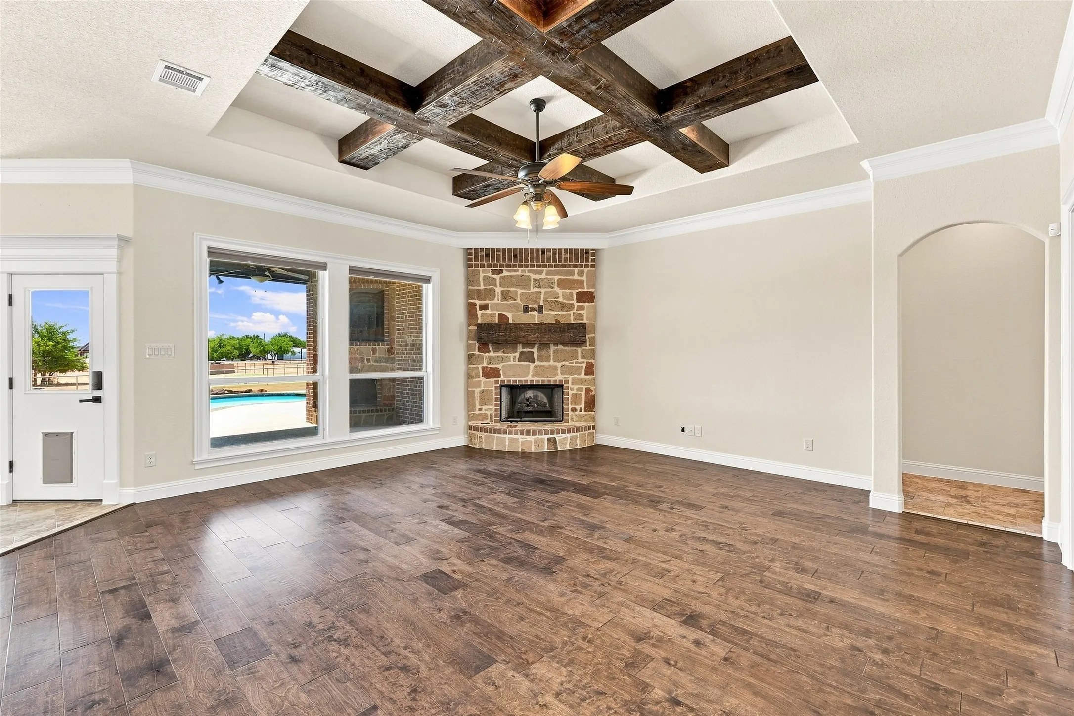 Vaulted ceiling with exposed wood beams and a ceiling fan, stone-clad corner fireplace with a wood mantel, wide plank wood-finish flooring, and large windows