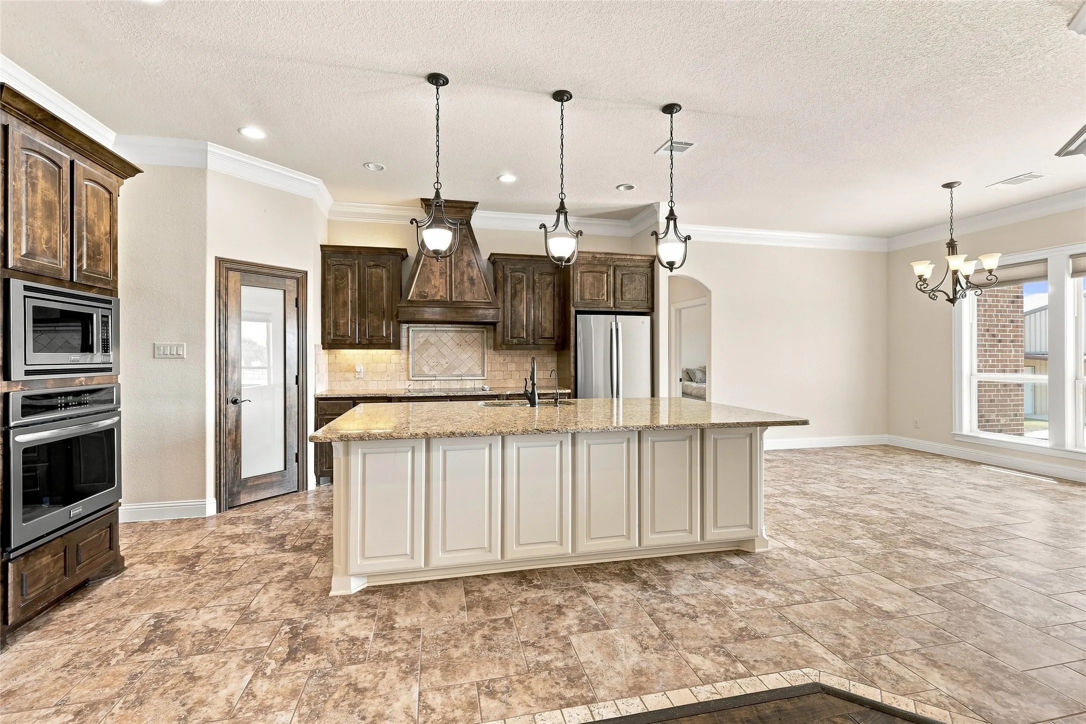 Spacious kitchen featuring an island with stone countertops, built-in ovens and microwave, dark wood-finish cabinetry, and tile flooring