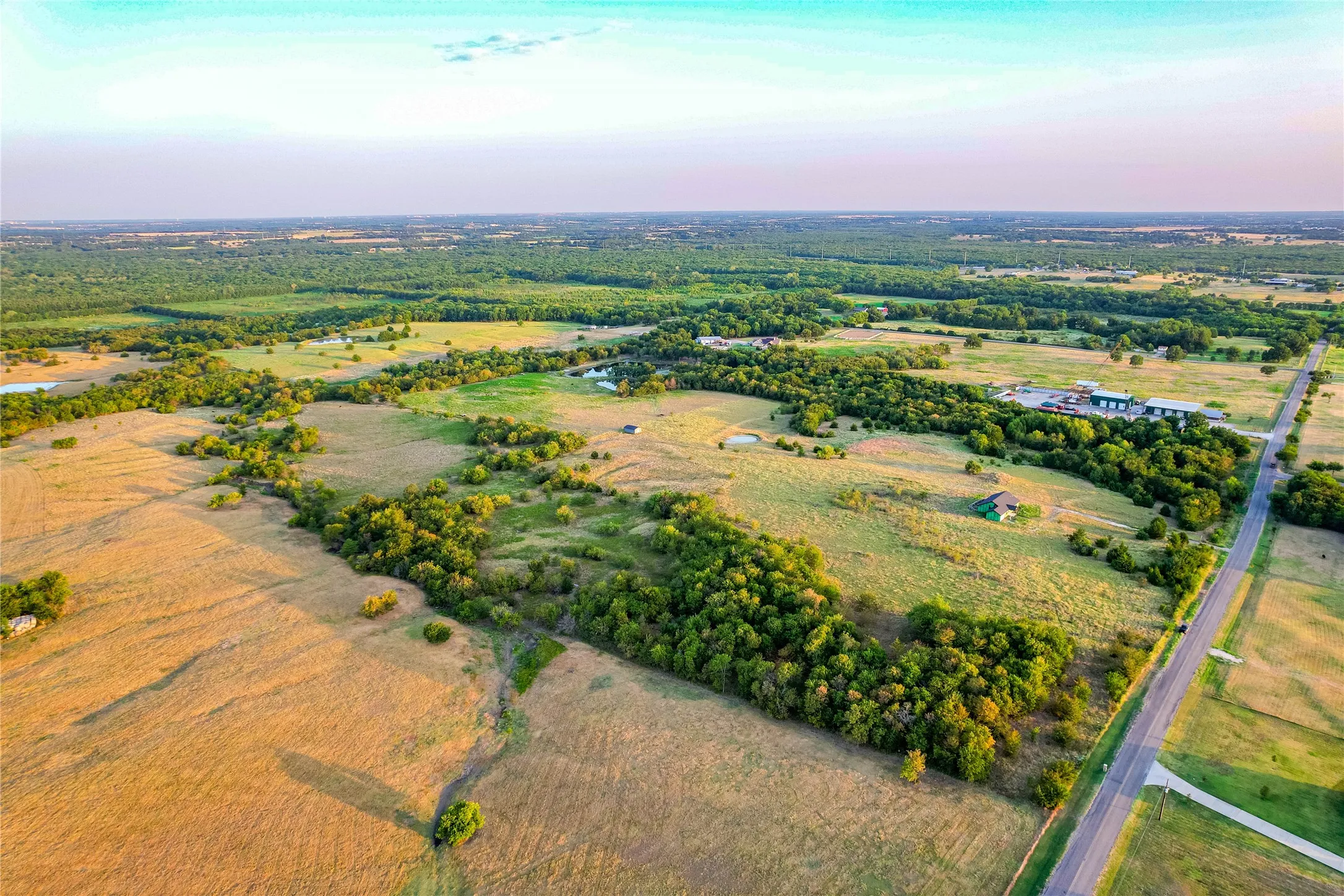 Expansive acreage featuring a paved road, scattered mature trees, open fields, and distant structures