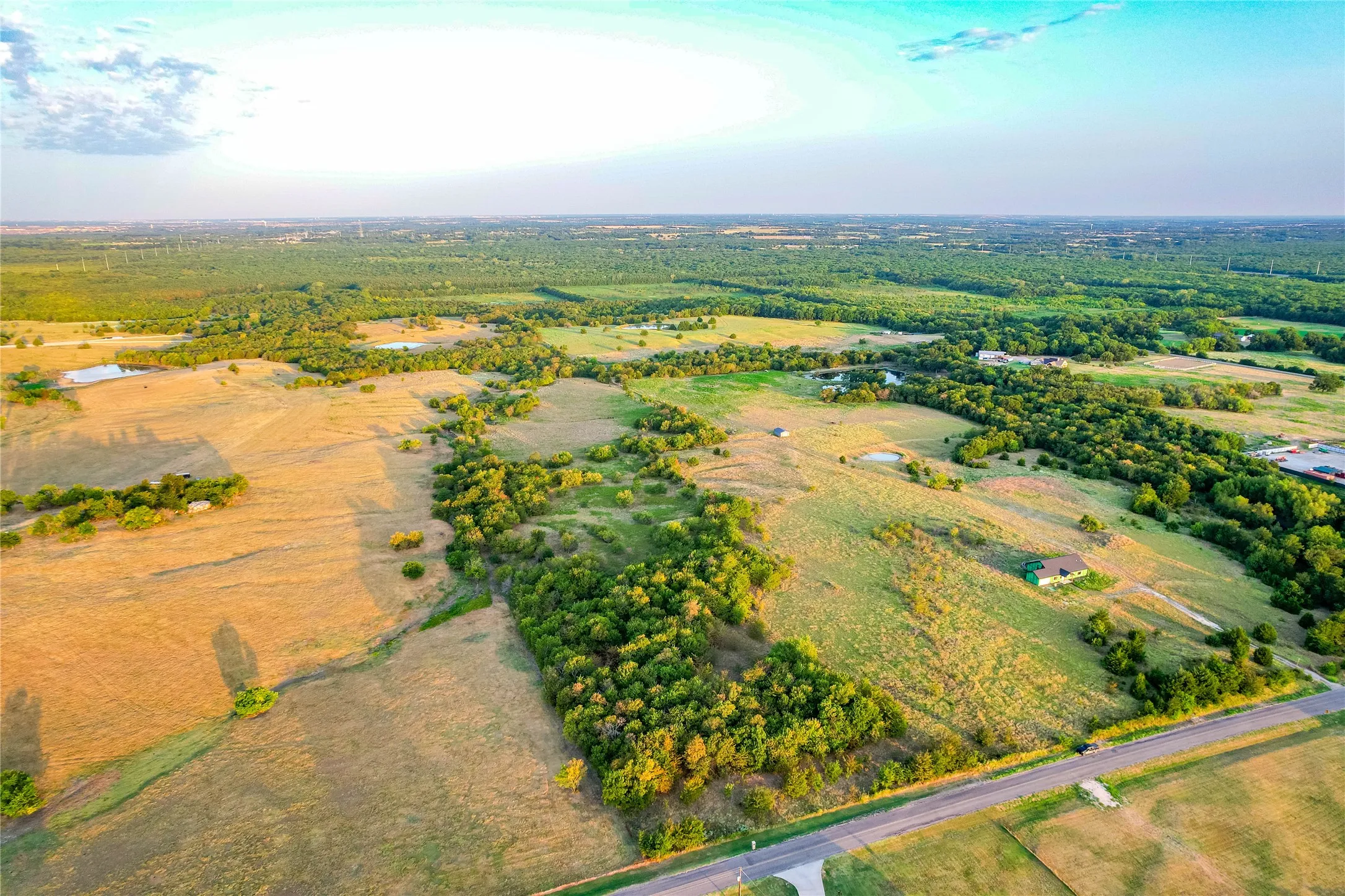 Expansive acreage featuring a paved road, scattered mature trees, and a distant structure