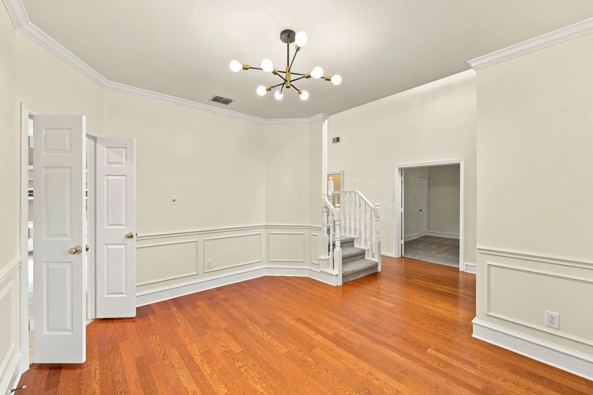 Dining area featuring rich wood-finish flooring, wainscoting, crown molding, a contemporary chandelier, and a carpeted staircase.