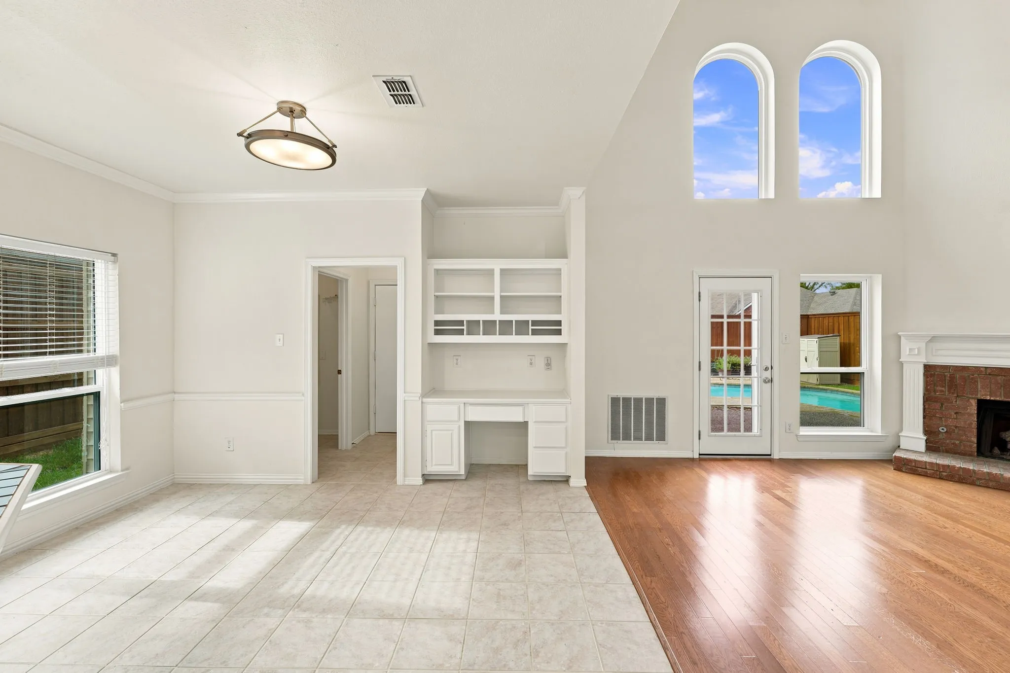 Two-story living area with high ceilings, featuring a brick fireplace with a white mantel, wood-finish flooring, and two arched windows