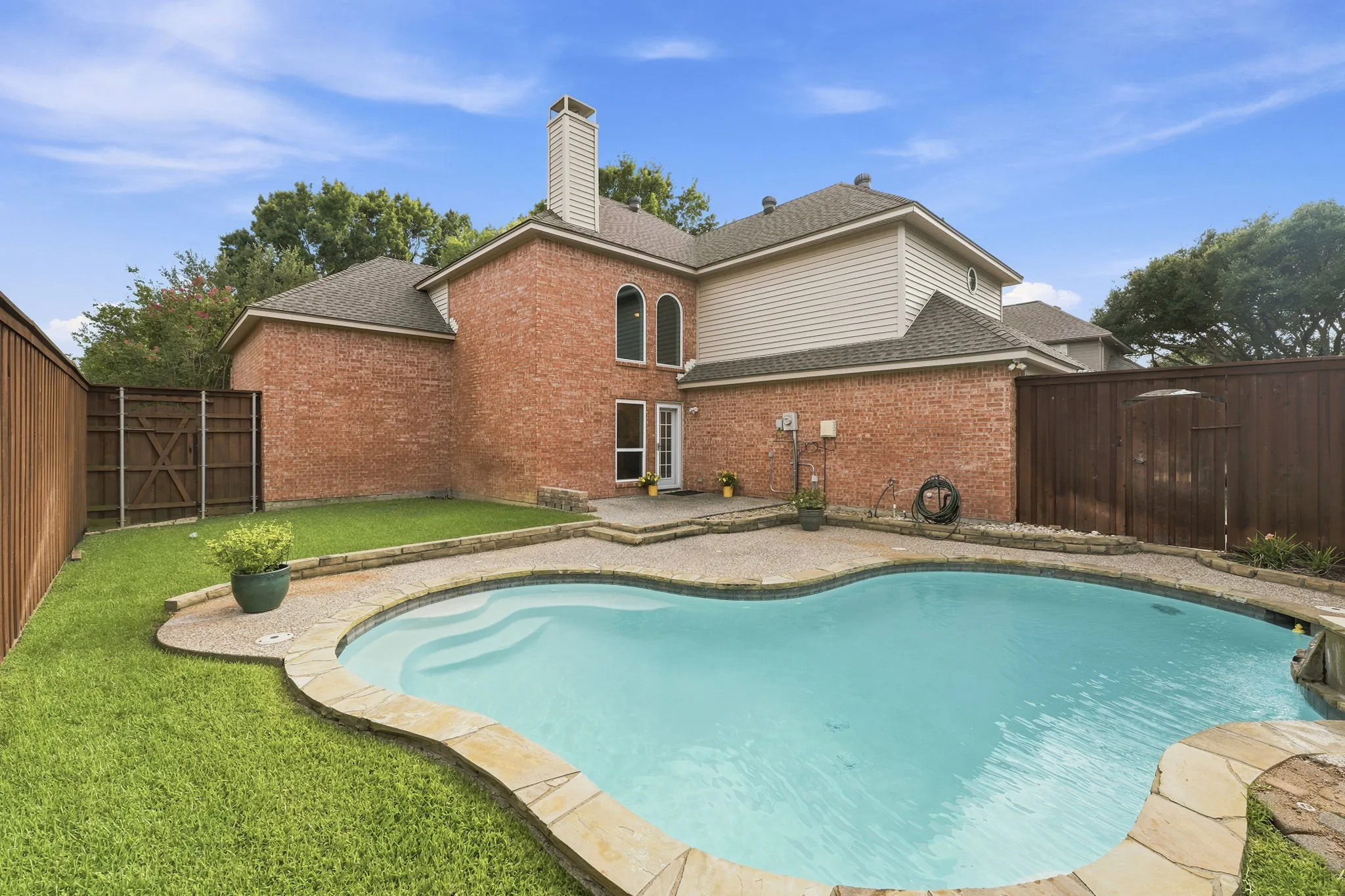 Brick exterior with siding accents, freeform swimming pool, stone coping, patio with brick pavers, and a wood privacy fence