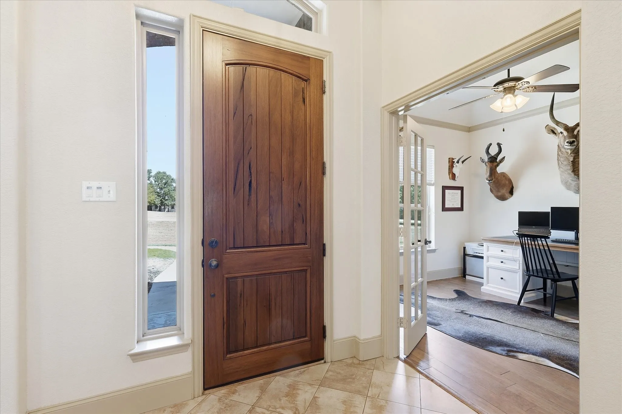 Arched wood-panel entry door with sidelights, tile flooring, and french doors opening to a room with wood-finish flooring and a ceiling fan