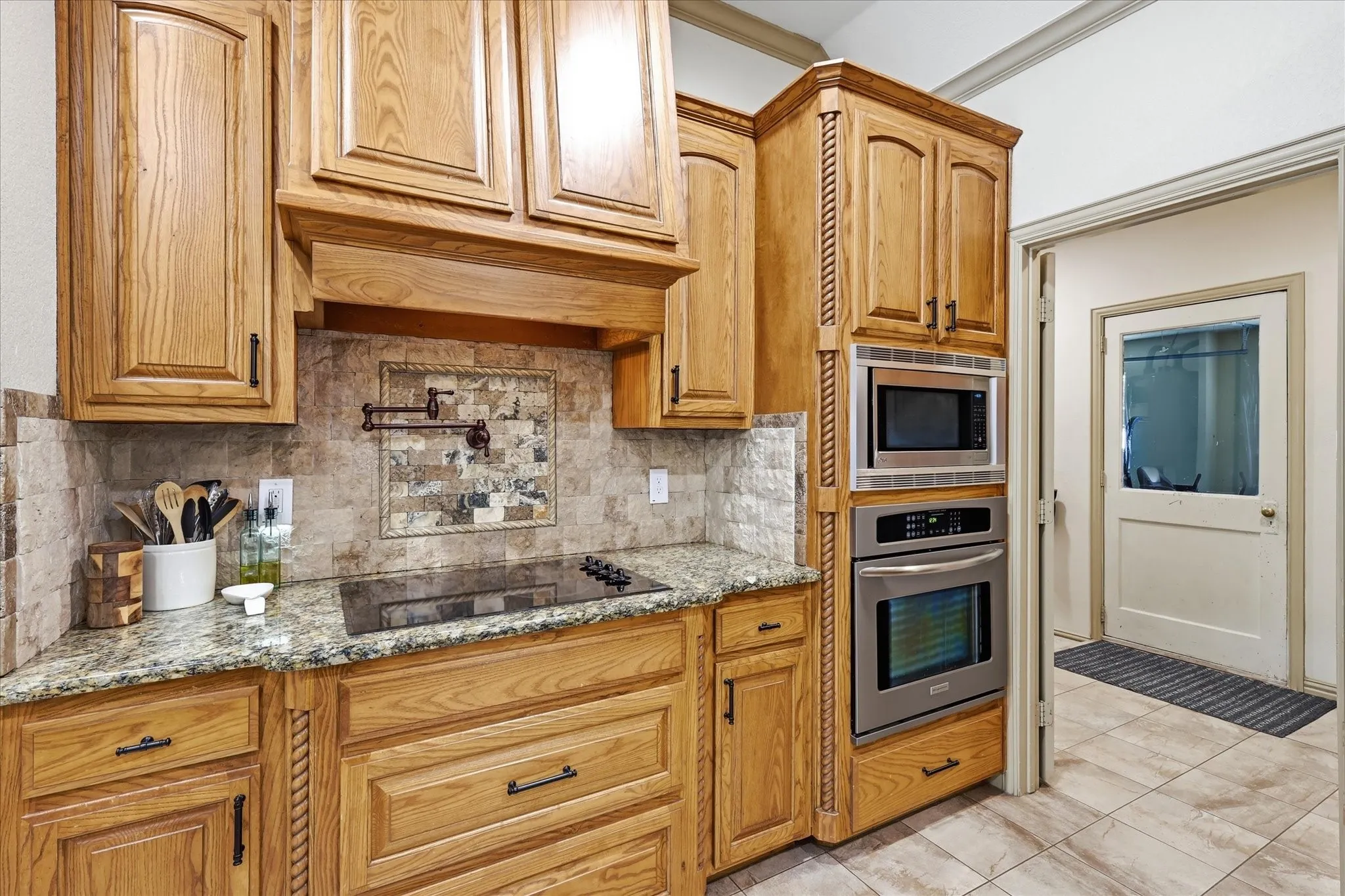 Wood cabinetry with decorative trim, granite countertops, tile backsplash with a decorative insert, stainless steel built-in appliances, and a smooth cooktop