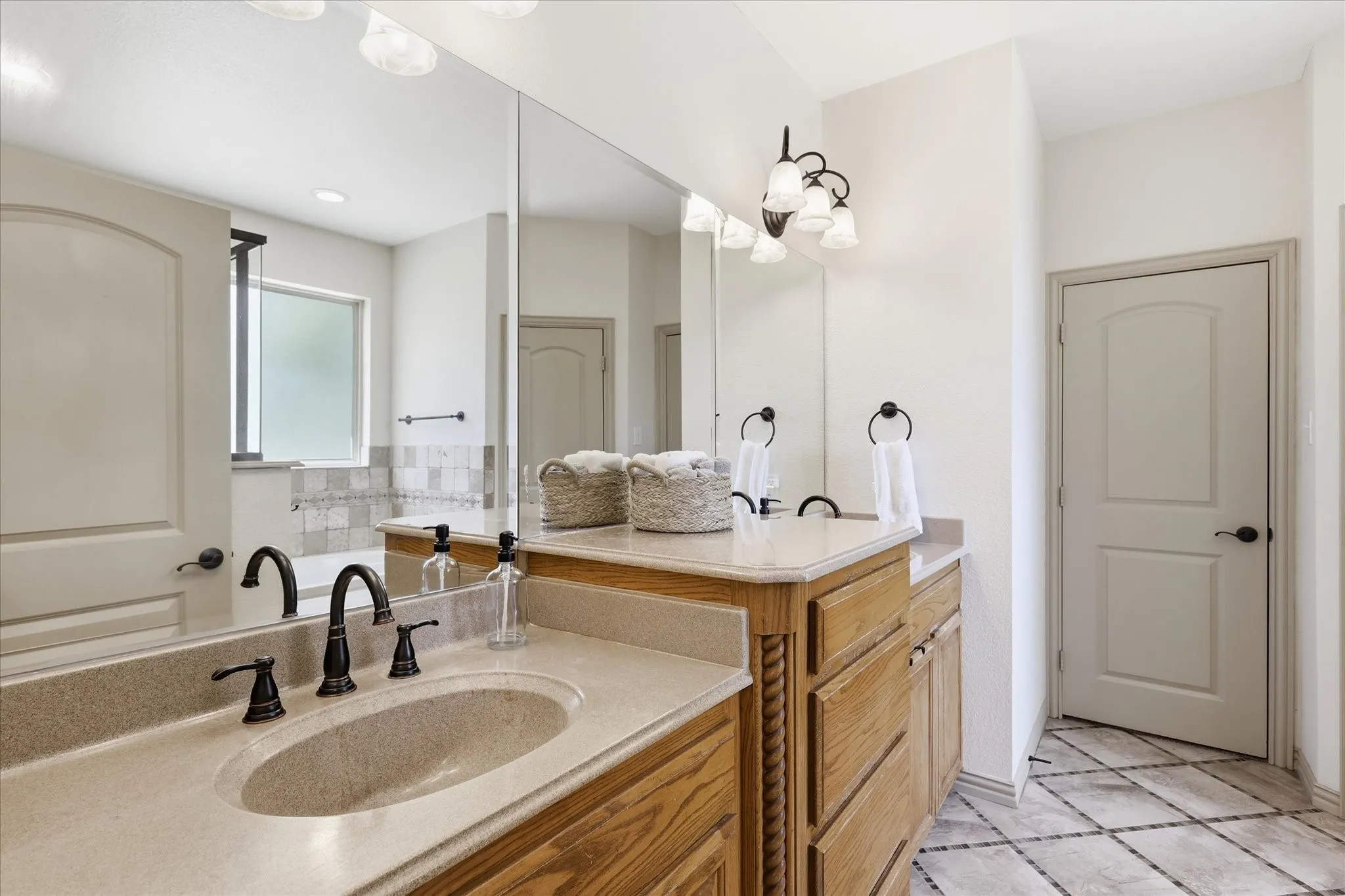Dual vanity with integrated sinks and bronze-finish fixtures, wood-finish cabinetry, full-width mirror, and patterned floor tile