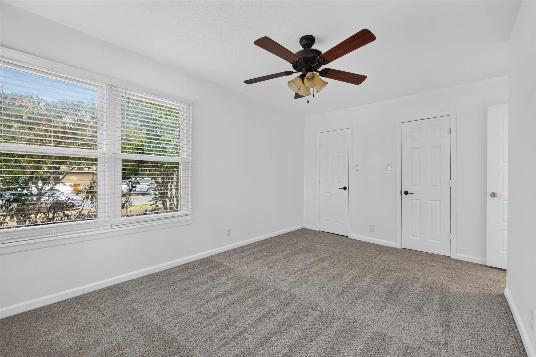 Dual windows with horizontal blinds, overhead ceiling fan with light fixture, gray carpeting, and white paneled doors