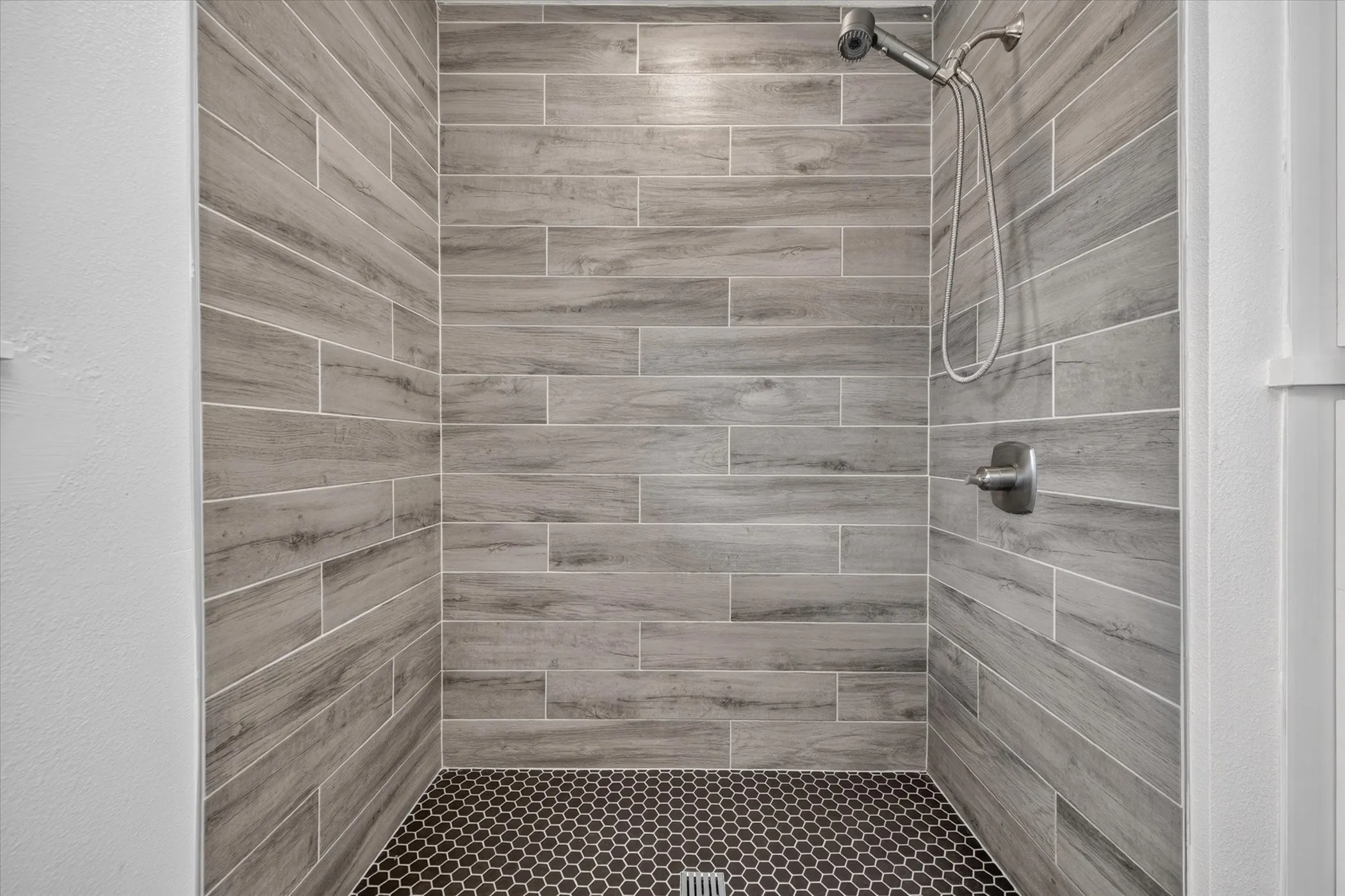 Shower stall with wood-finish wall tile, dark hexagonal floor tile, and brushed nickel fixtures
