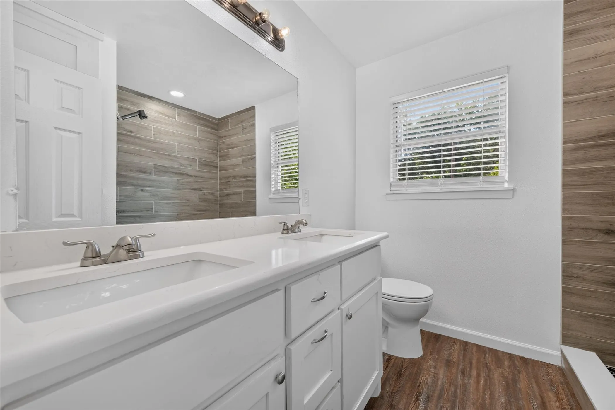 Dual vanity with integrated sinks, brushed nickel fixtures, wood-finish flooring, and a tiled shower surround
