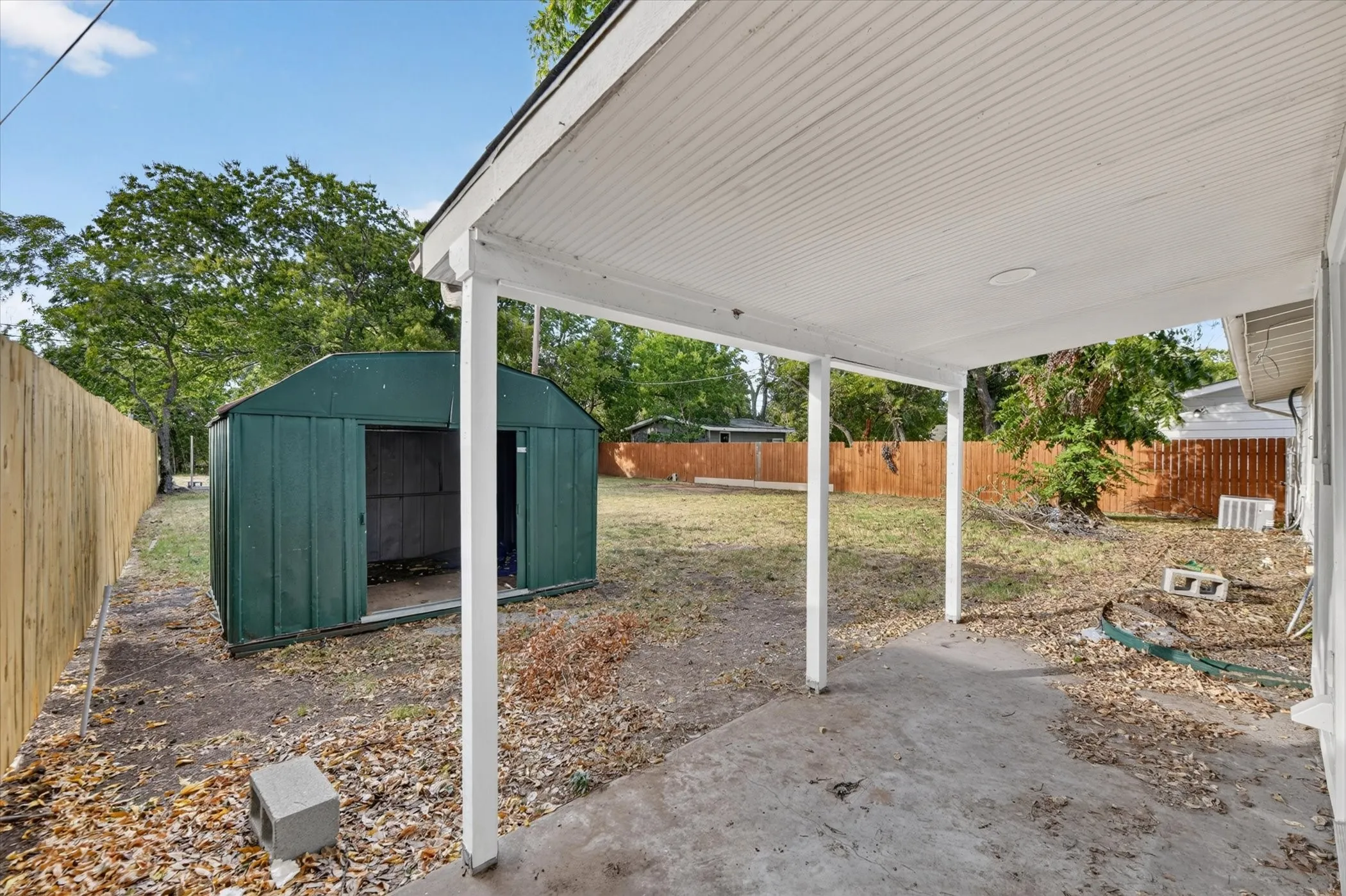 Covered concrete patio with white support columns, privacy fencing, and a detached storage shed