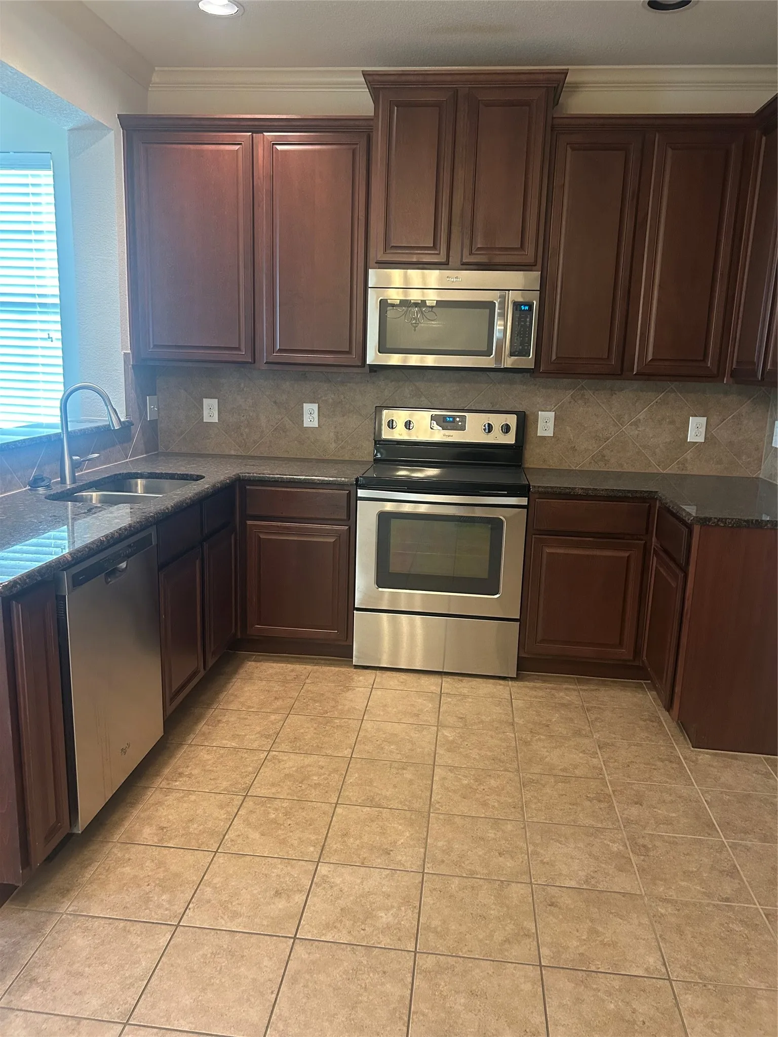 Kitchen featuring dark wood-finish cabinetry, stainless steel appliances, dark granite countertops, tile backsplash, and tile flooring