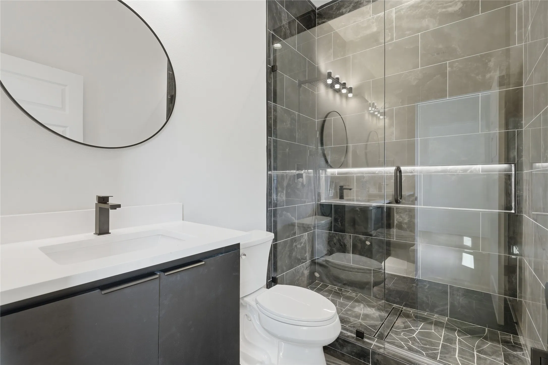 Frameless glass shower enclosure, dark tiled shower surround, integrated shower niche, dark wood-finish vanity with a white countertop, and a circular black-framed mirror