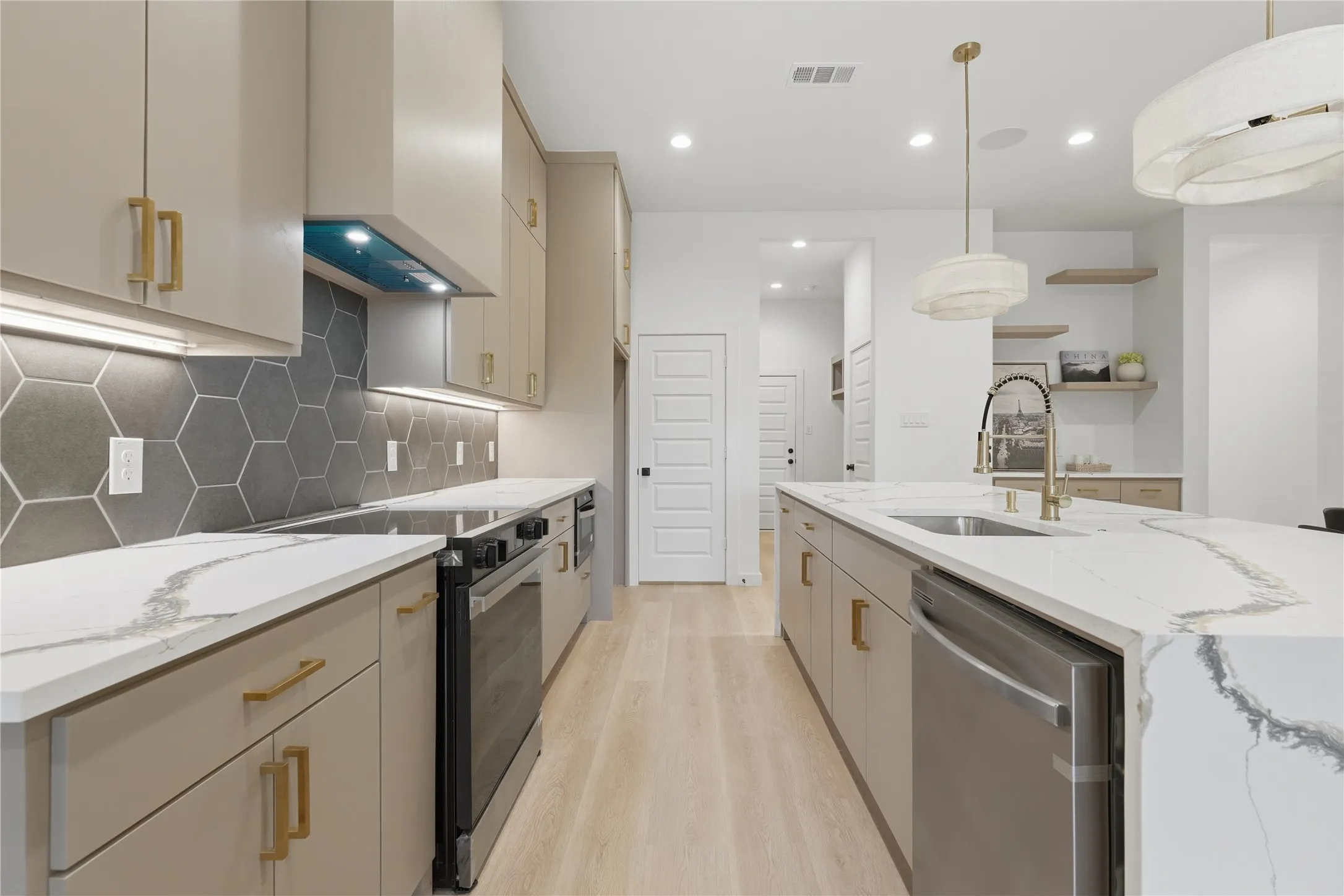 Modern kitchen featuring light cabinetry, a large center island with integrated sink, stainless steel appliances, hexagon tile backsplash, and light wood-finish flooring