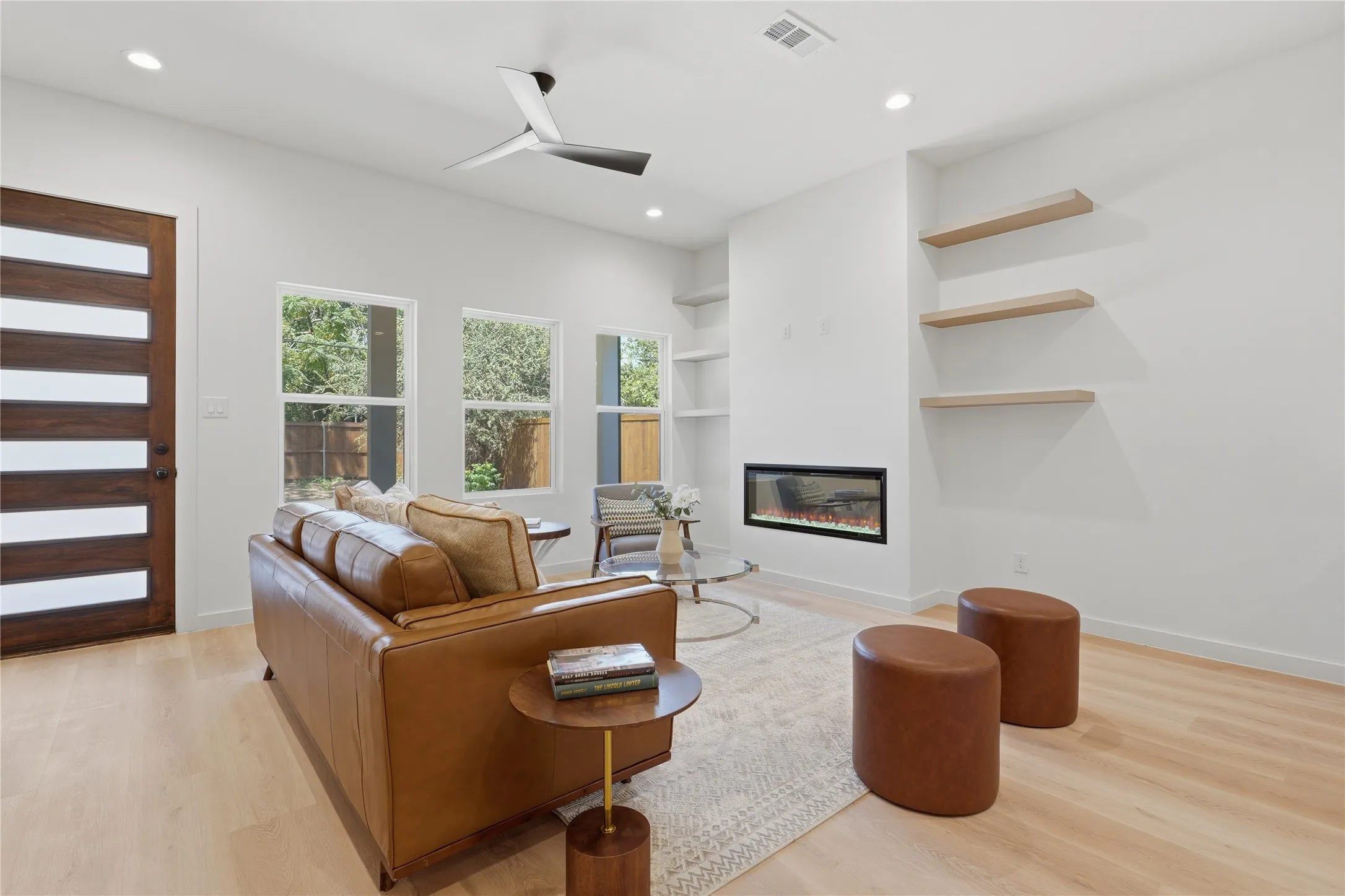 Recessed lighting, wood-finish flooring, a modern ceiling fan, three windows with exterior views, and a built-in electric fireplace with floating shelves