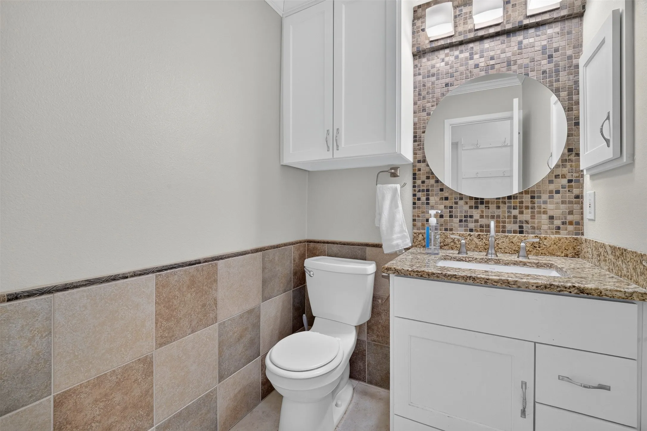White vanity with a granite-style countertop, mosaic tile backsplash, and a round mirror