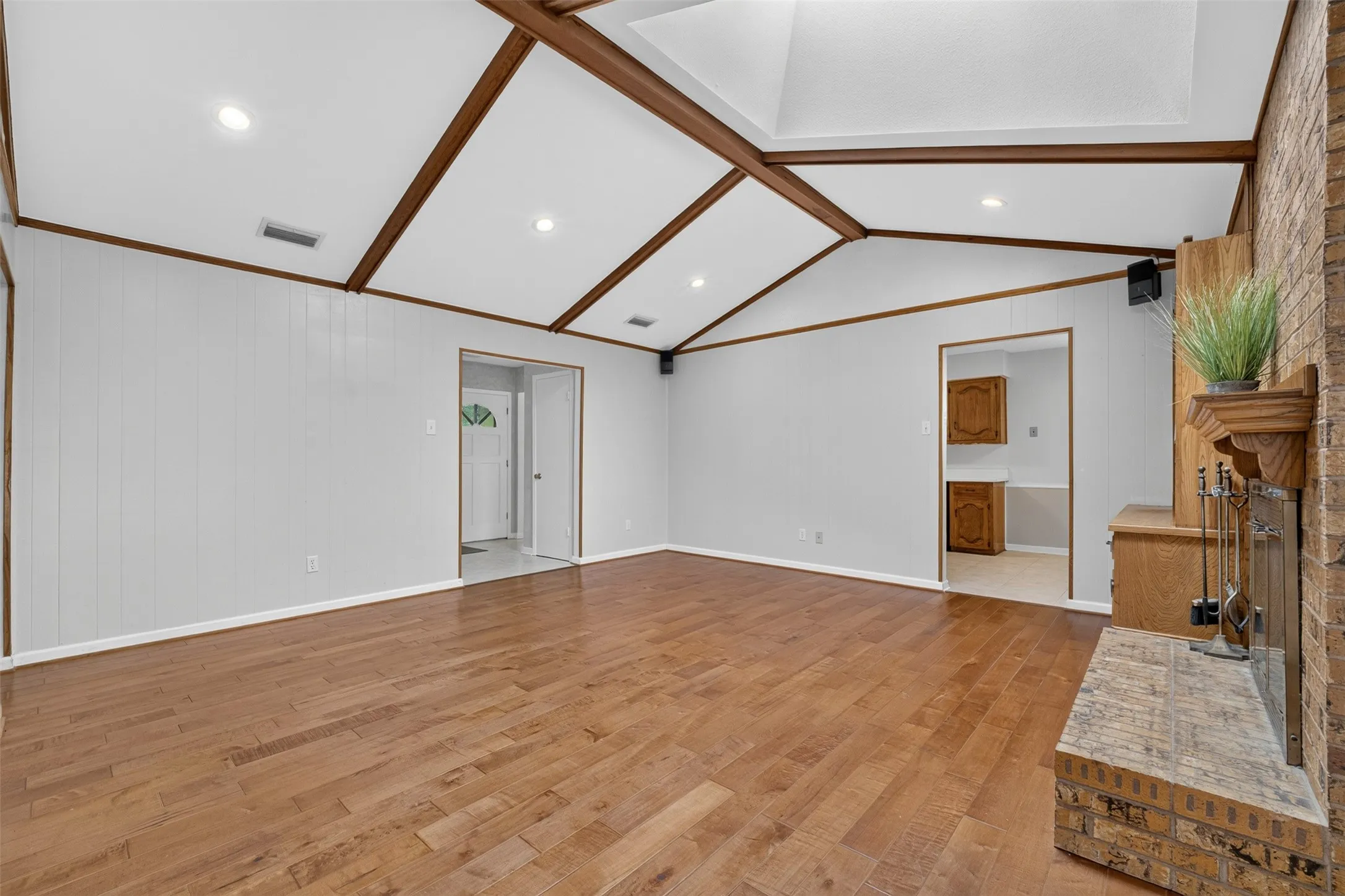 Vaulted ceiling with exposed wood beams, recessed lighting, wood-finish flooring, brick fireplace with mantel, and built-in shelving