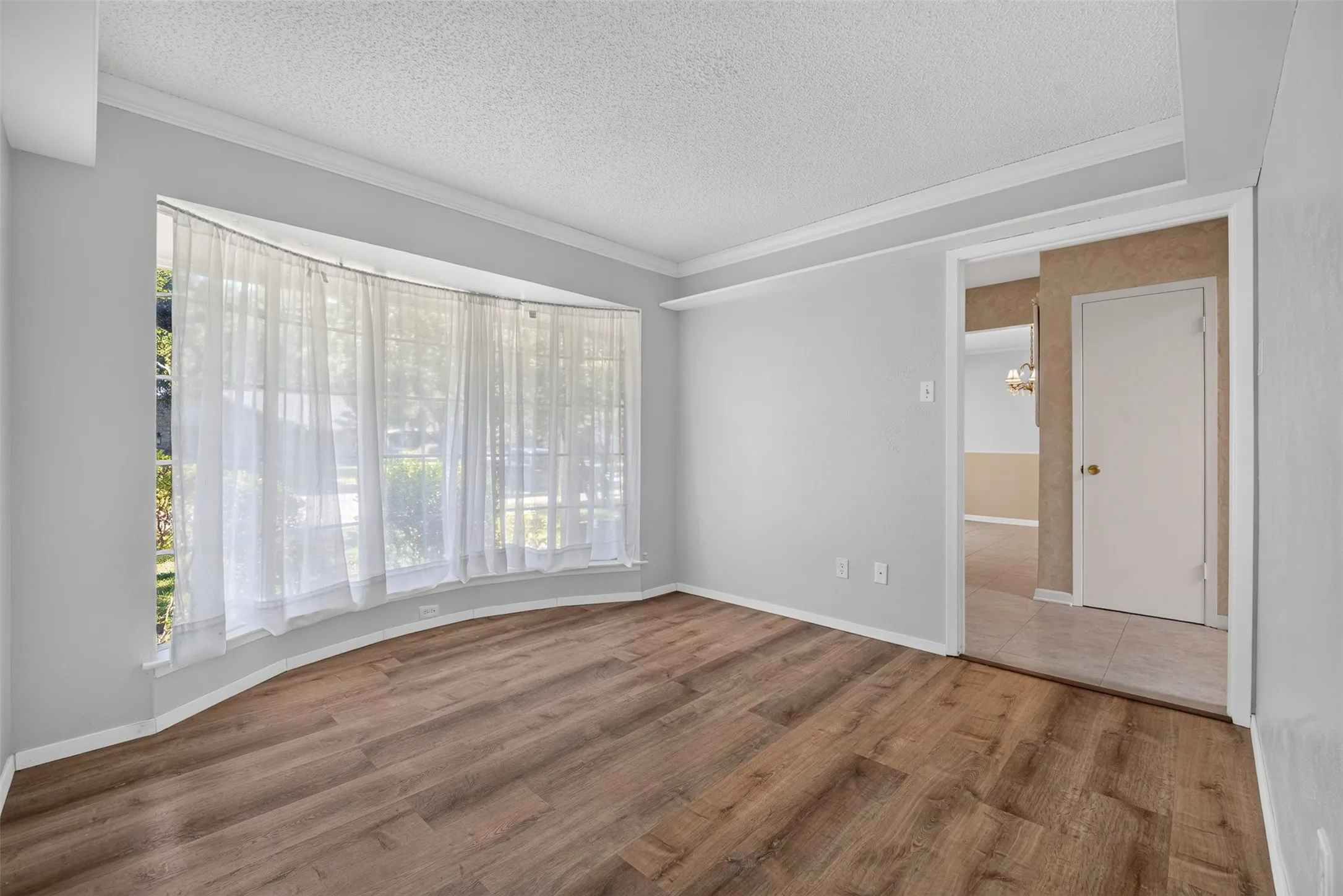 Bay window with sheer curtains, wood-finish flooring, light gray wall paint, and white crown molding