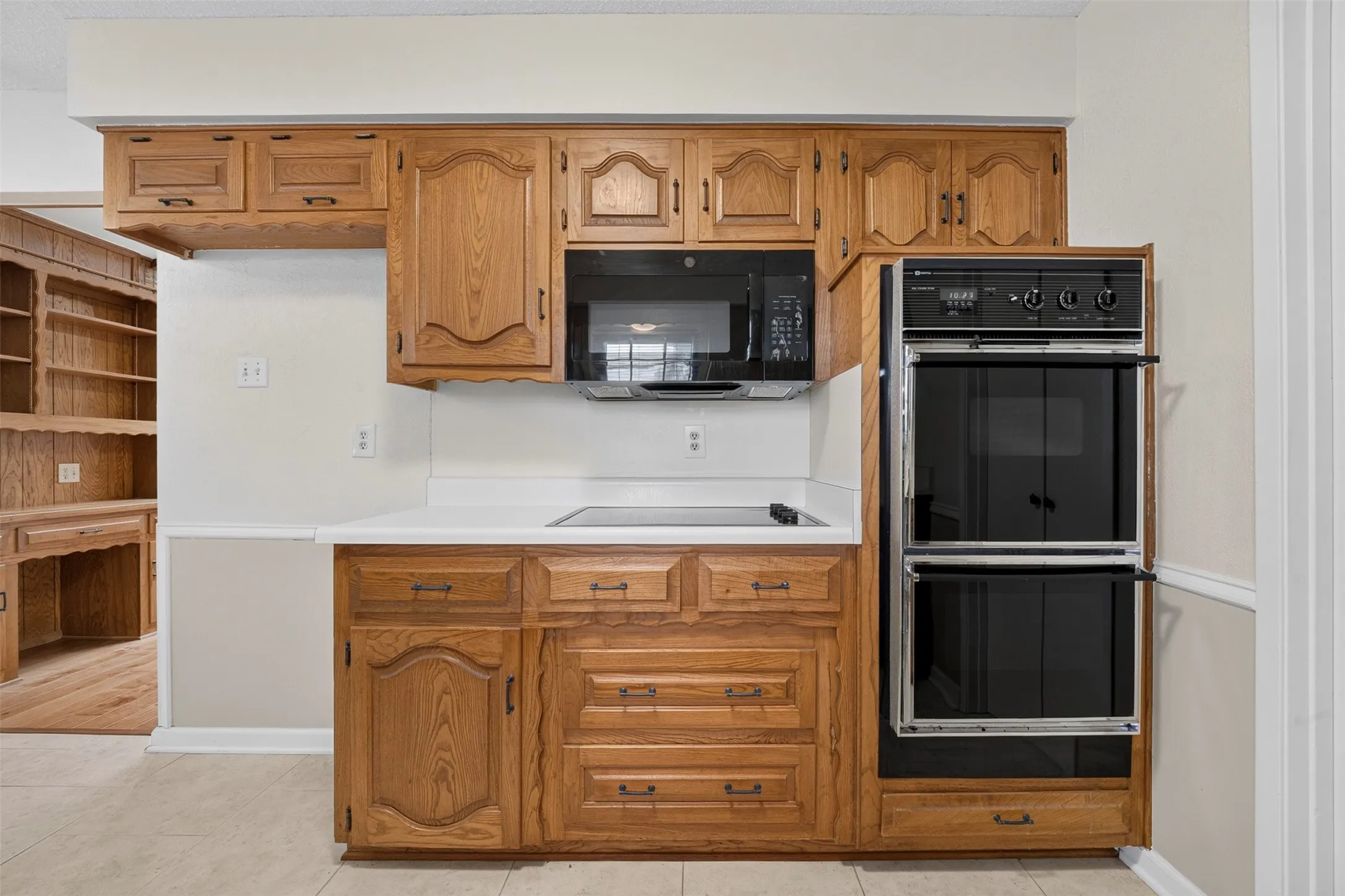 Wood-finish cabinetry with dark hardware, white countertops, built-in microwave, double wall oven, and ceramic tile flooring