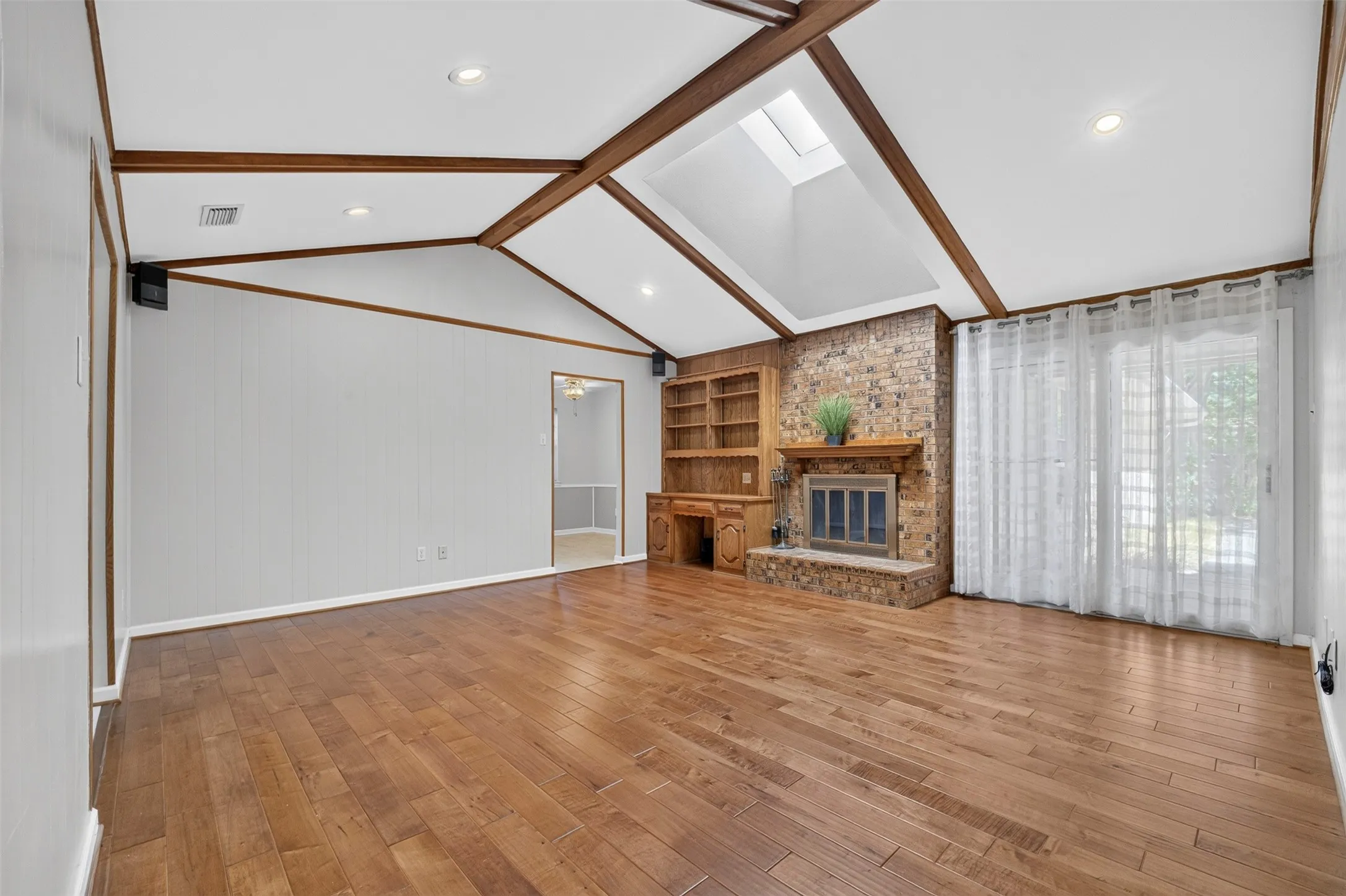 Vaulted ceiling with exposed wood beams, wood-finish flooring, brick-surround fireplace with hearth, built-in shelving, and a large skylight