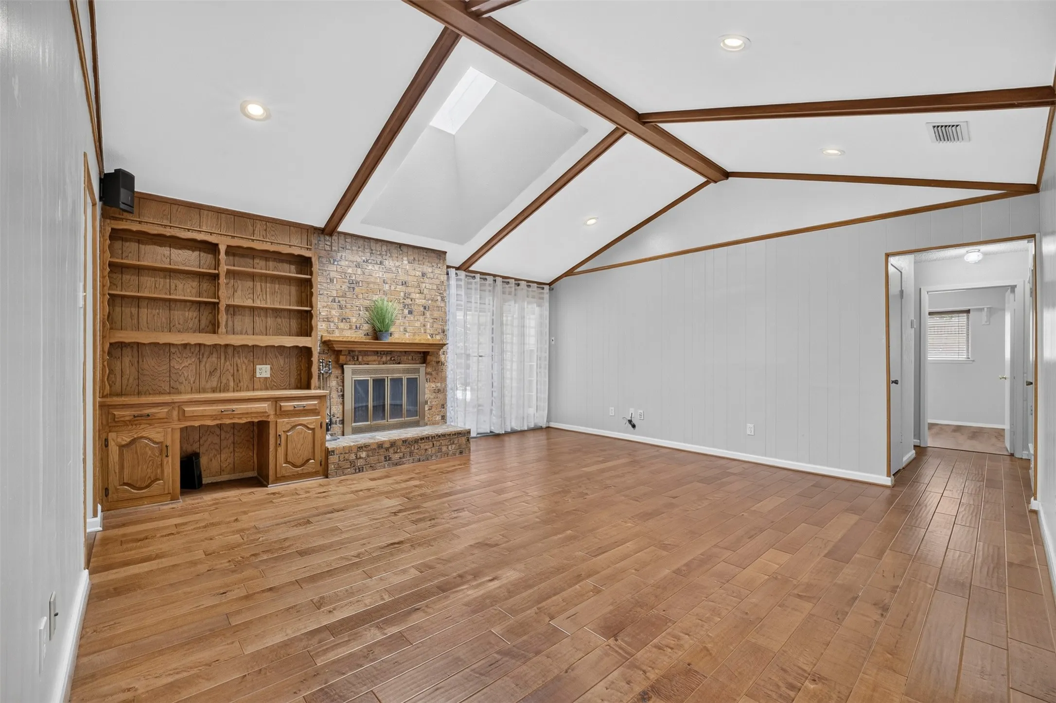 Vaulted ceiling with exposed beams, wood-finish flooring, stone-clad fireplace, built-in wooden shelving and cabinetry, and a skylight