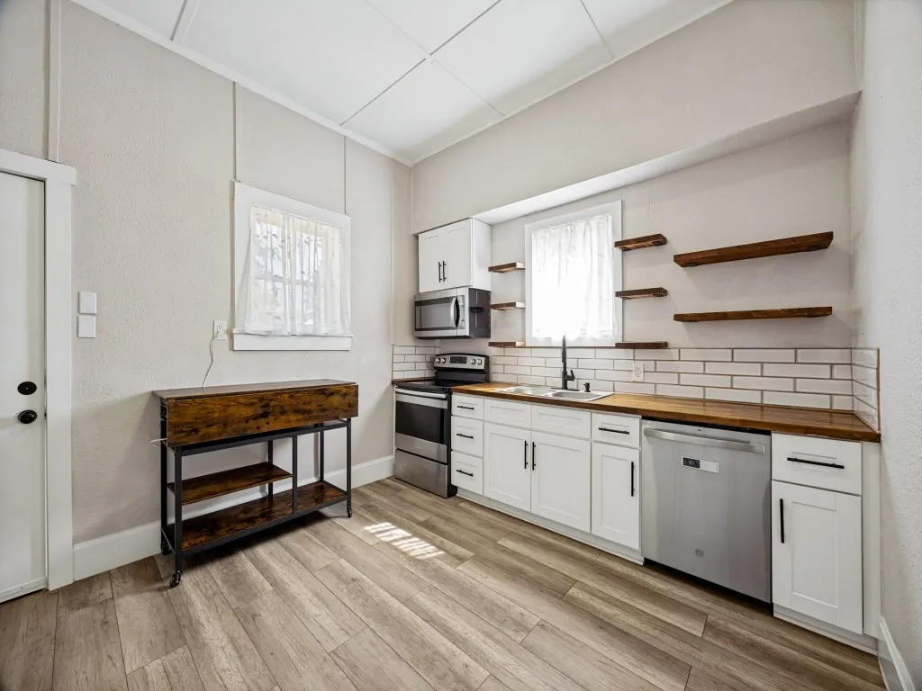 White cabinetry, rolling island, butcherblock wood-finish countertops, subway tile backsplash, wood-finish flooring, and stainless steel appliances