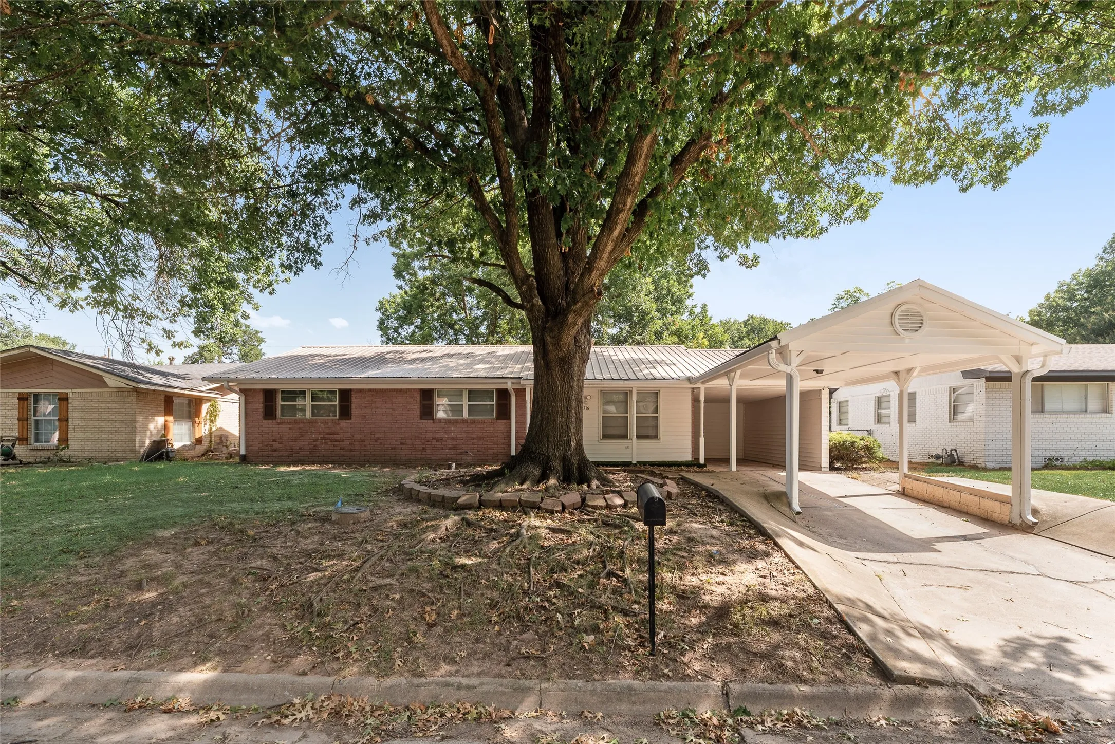 Exterior featuring a white carport, mature shade tree, brick facade, and a metal roof