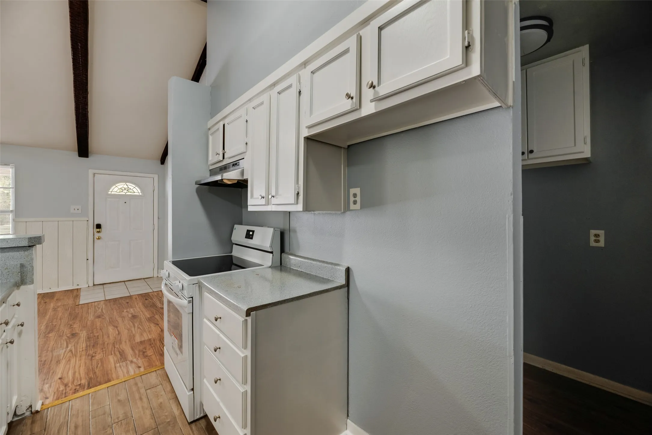 Vaulted ceiling with exposed wood beams, wood-finish flooring, white cabinetry, integrated range, and stone-finish countertops