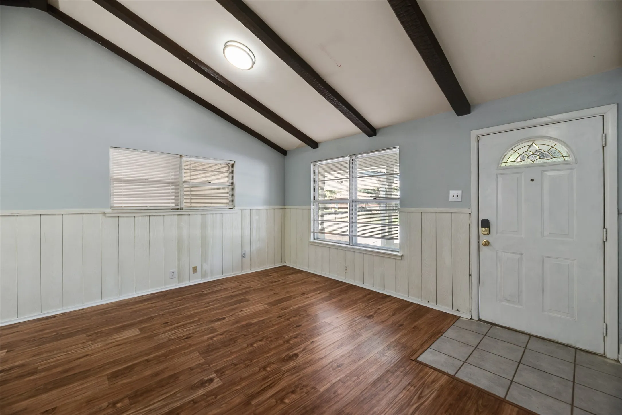 Vaulted ceiling with exposed beams, wood-finish flooring, white paneled wainscoting, and a front entry door with a decorative glass insert