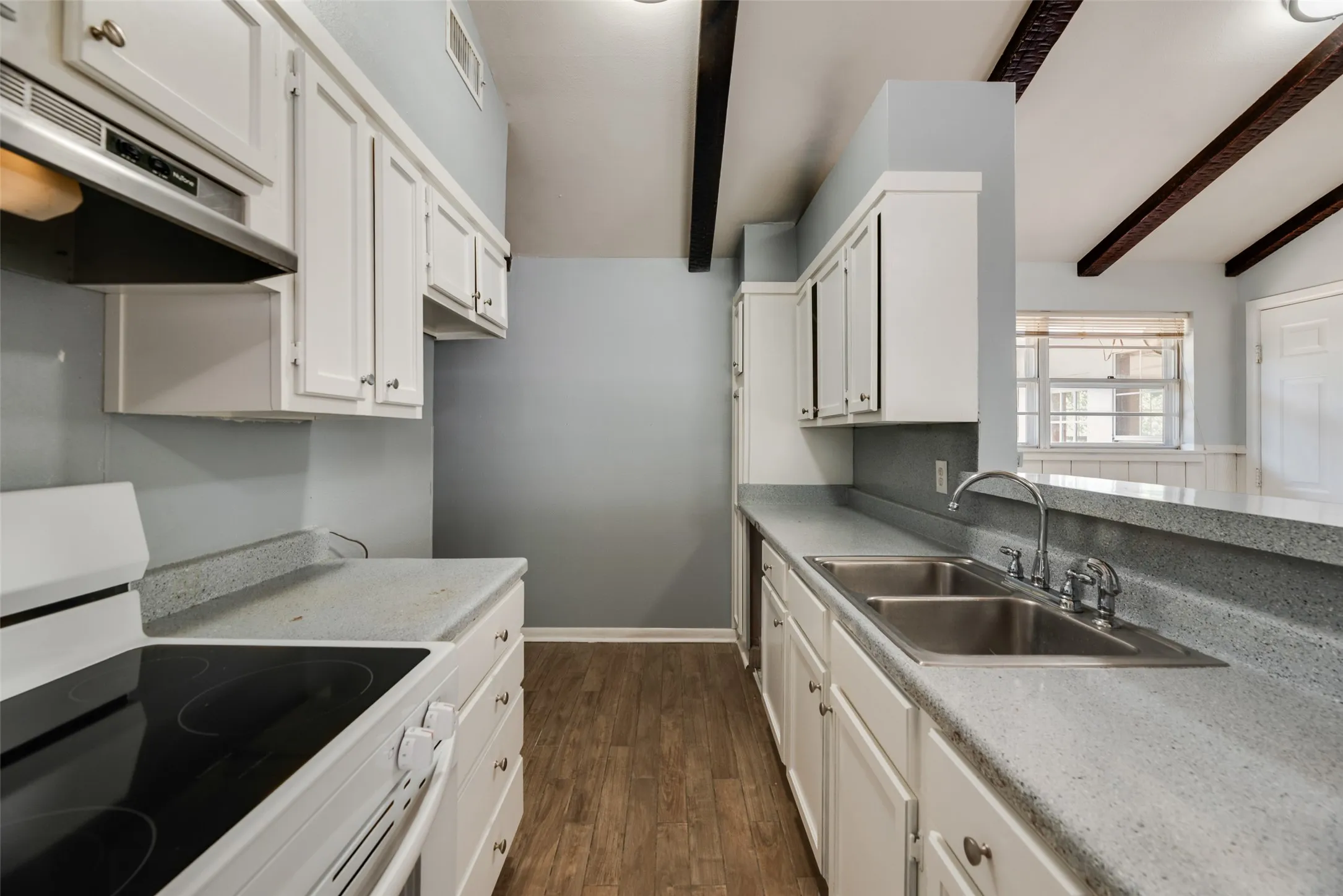 Galley kitchen featuring white cabinetry, gray countertops, a double basin stainless steel sink, wood-finish flooring, and exposed ceiling beams