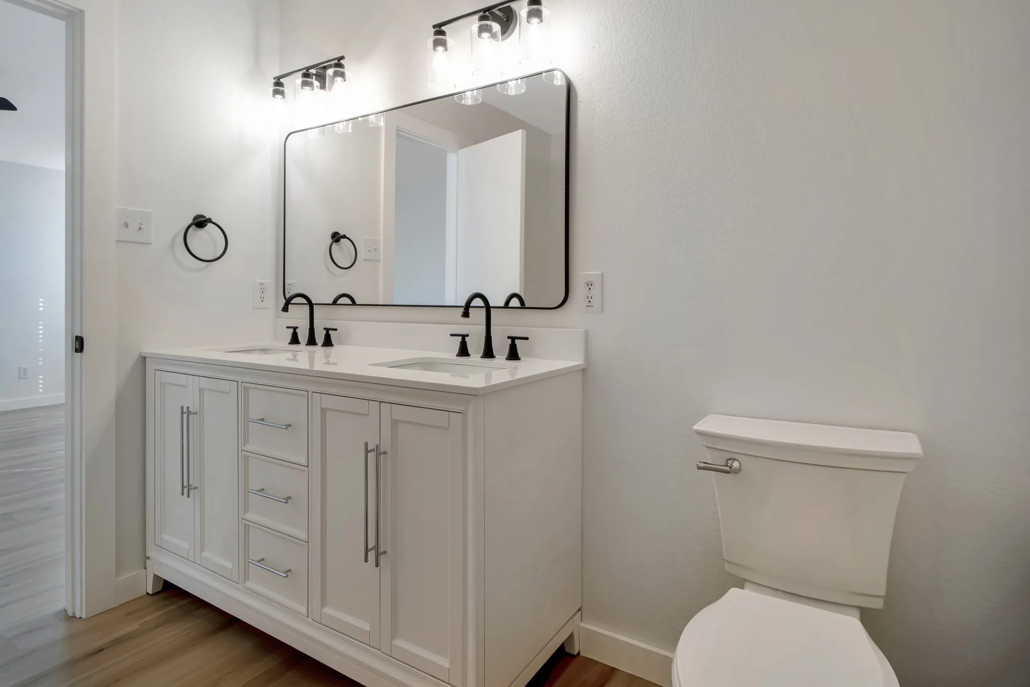 Dual vanity with white cabinetry and integrated sinks, matte black hardware, large rectangular mirror, and wood-finish flooring