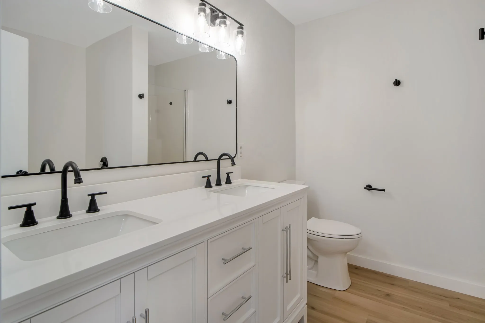 Dual vanity with white cabinetry and integrated sinks, matte black hardware, large rectangular mirror, and wood-finish flooring