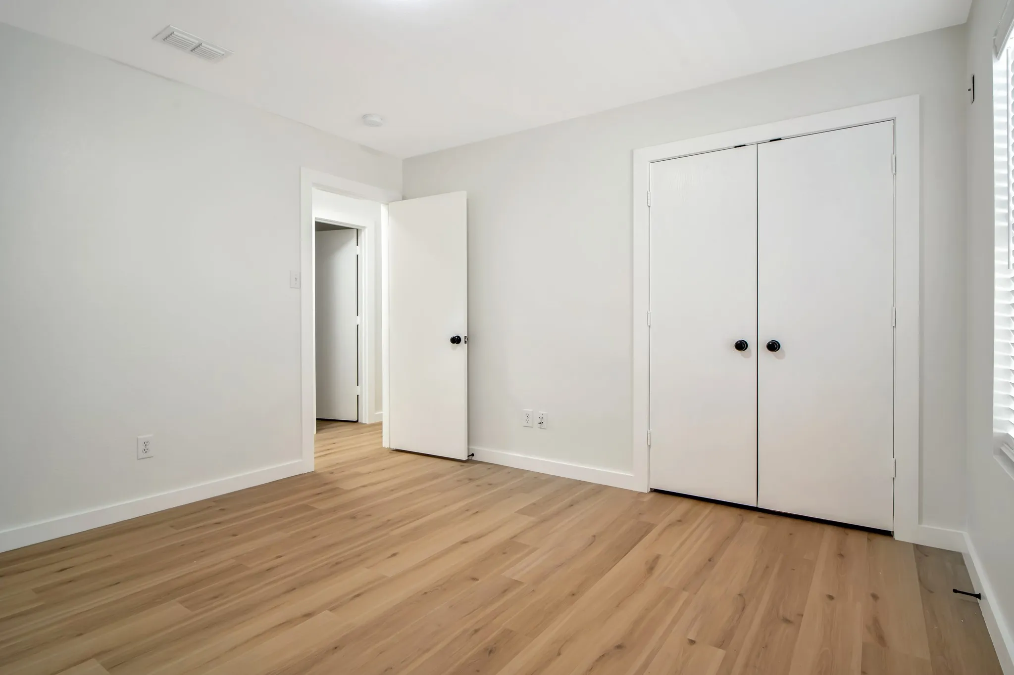 Light-toned wood-finish flooring, white walls, white bi-fold closet doors, and a window with white blinds