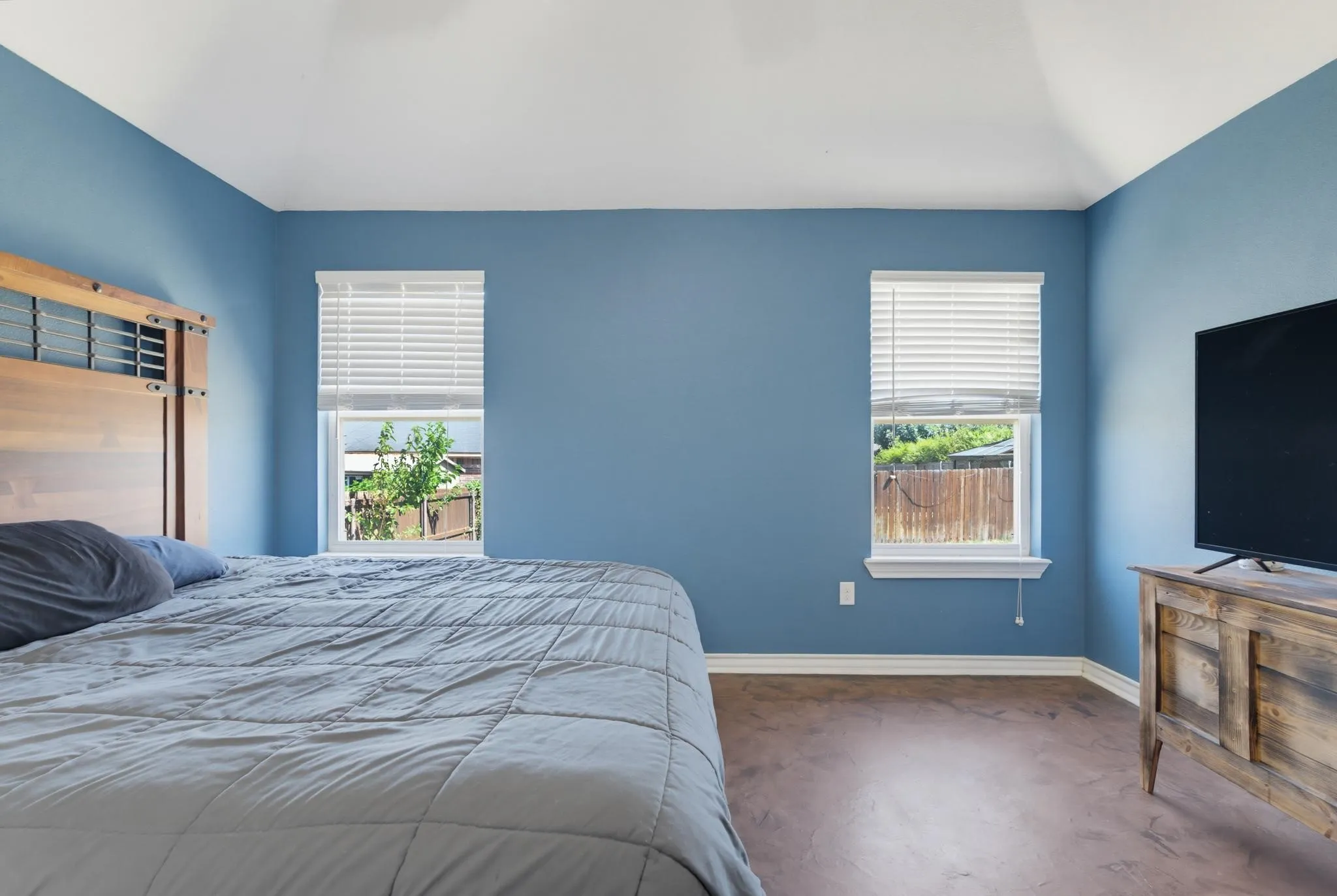 Serene primary bedroom showcasing calming blue walls, abundant natural light from dual windows with plantation shutters, wood-finish flooring, and a wooden dresser with integrated television mount